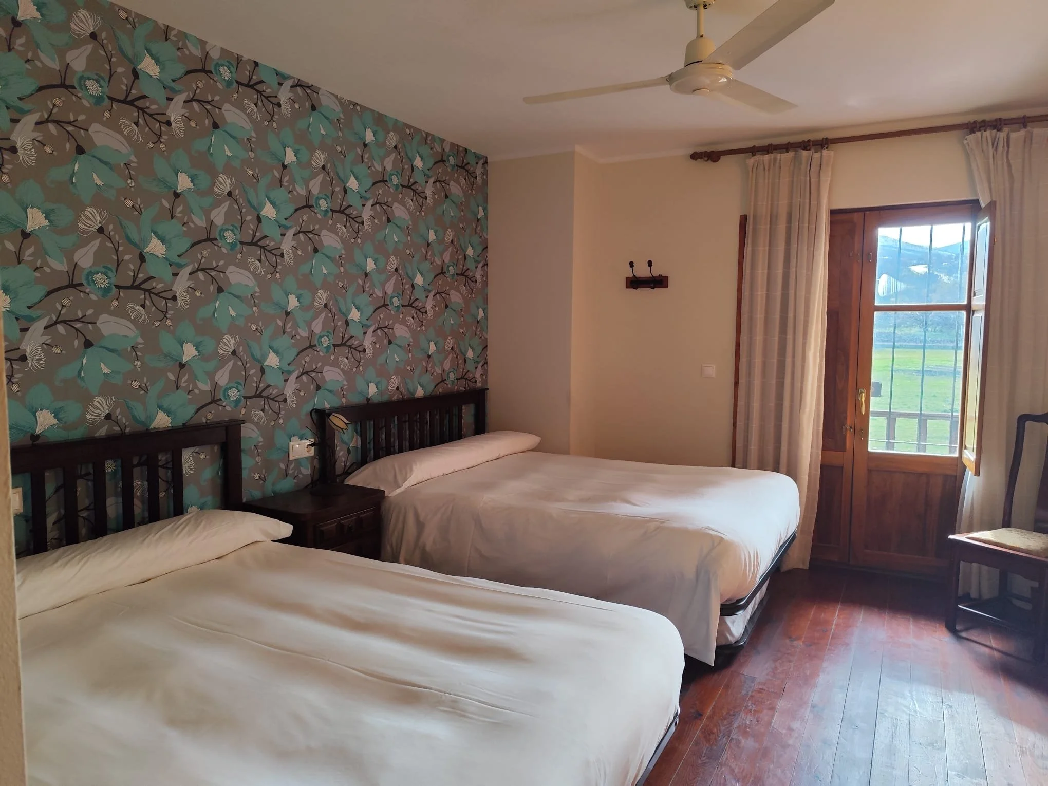 Habitación con dos camas de 1,35 en hotel rural la peregrina con cabeceras de madera, una pared con papel tapiz con diseño de ramas y hojas en tonos azules y beige, ventana de madera con cortinas claras, suelo de madera y ventilador en el techo.