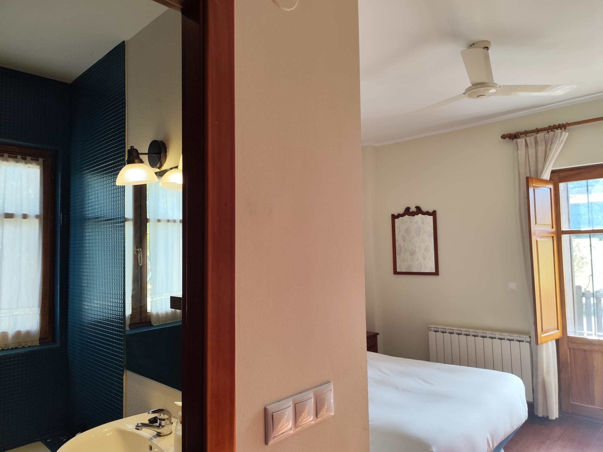 Imagen de una habitación de hotel con cama, ventanas con cortinas blancas y puertas abiertas, vista hacia un área con paredes blancas y una luz blanca en la pared.