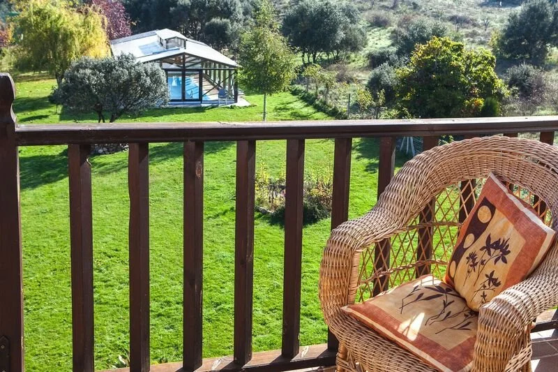 Vista de un patio con césped, varios árboles y una estructura de vidrio en el fondo. En el balcón, una silla de mimbre con cojín decorativo.
