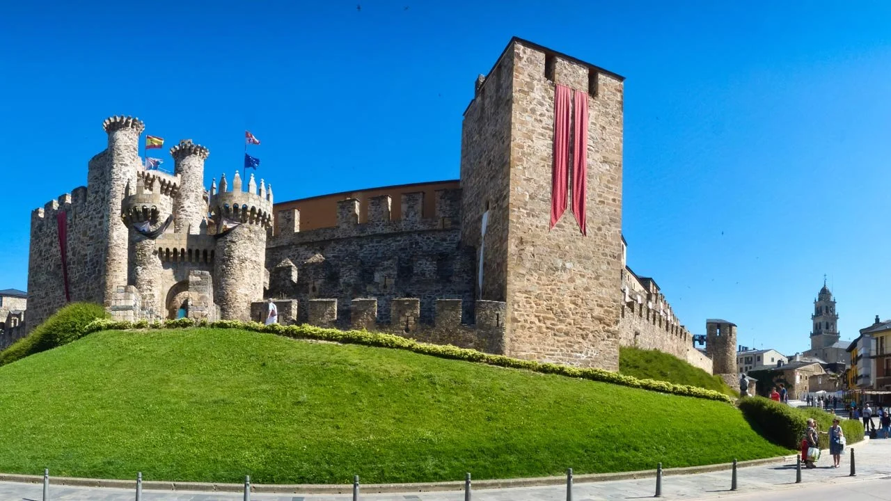 Castillo medieval de piedra con torres y murallas, en un día soleado, con una colina verde y un cielo azul, en una zona urbana con personas caminando y edificios alrededor.