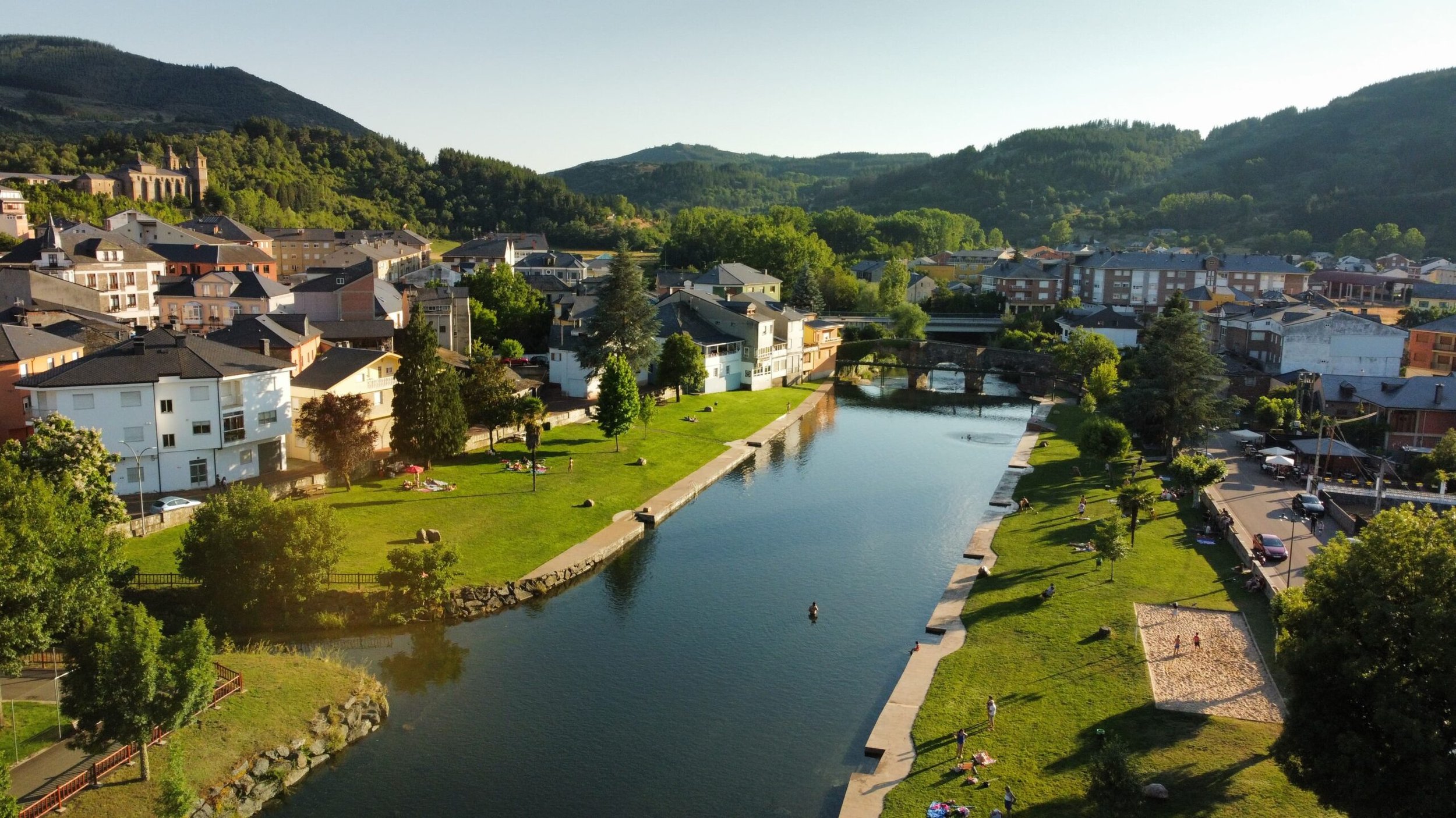 Vista aérea de un río en un parque urbano con árboles, césped y personas disfrutando, rodeado de casas y colinas verdes.