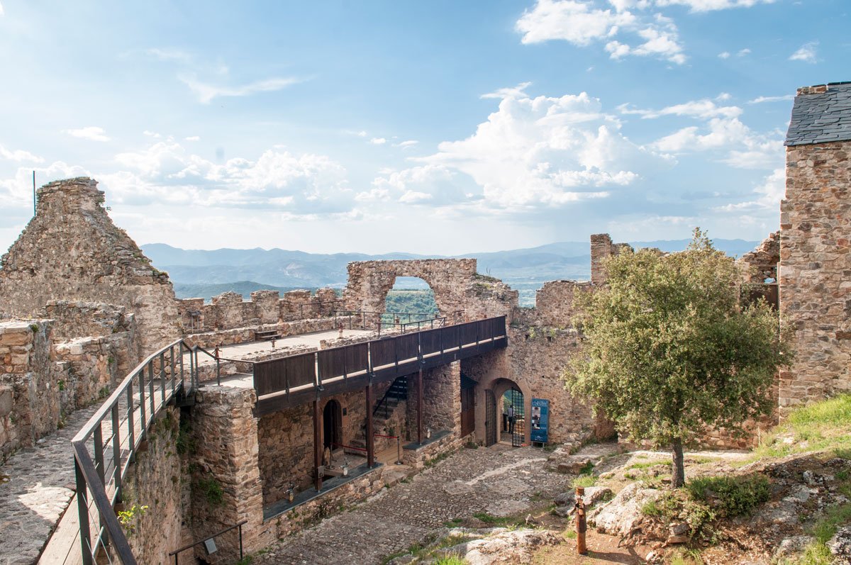 Restos de un castillo de piedra con un puente y un árbol en un paisaje con montañas al fondo y cielo con nubes.