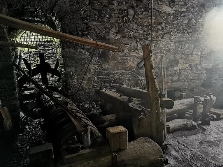 Interior de una antigua herrería con maquinaria de forjar, paredes de piedra y una pequeña ventana con rejas.