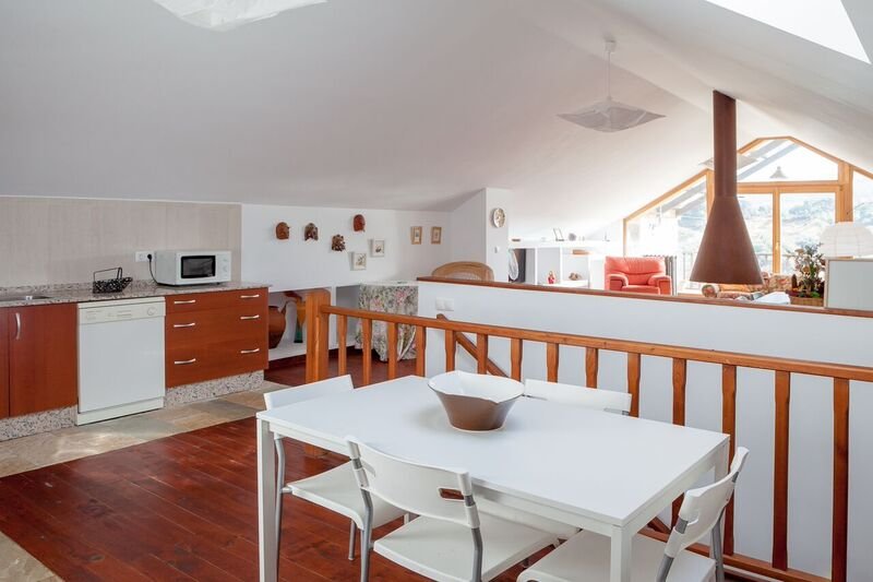 Interior de un apartamento con área de cocina, comedor y sala, con paredes blancas, muebles de madera y una gran ventana que permite entrada de luz natural.