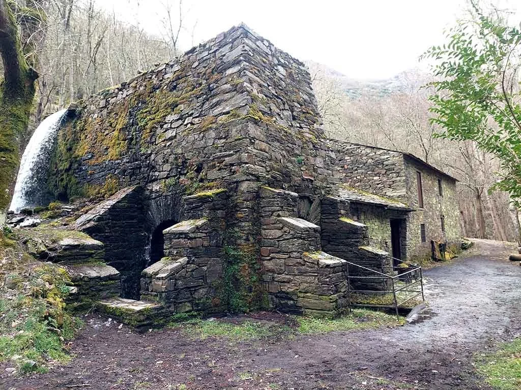 Antigua casa de piedra en medio del bosque, con una pequeña cascada en uno de sus lados y sendero de tierra al frente.