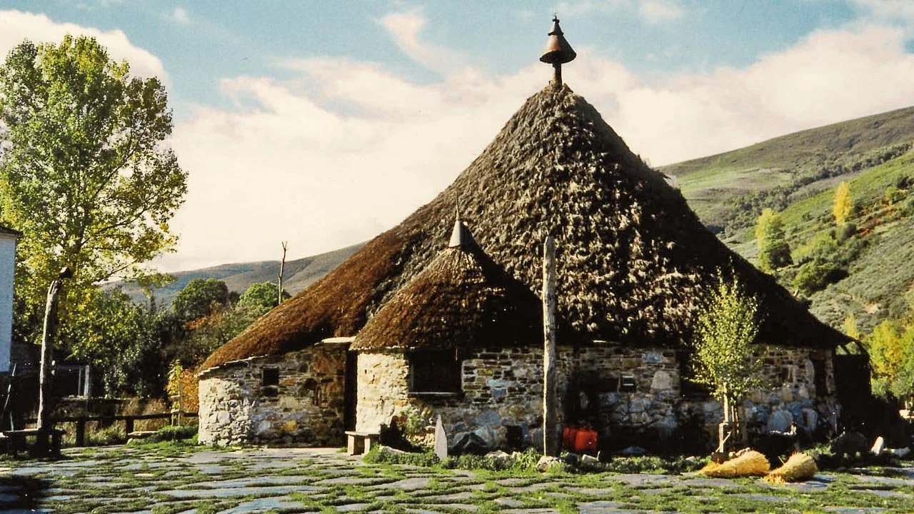 Casa tradicional de piedra con techo de paja en un paisaje rural con montañas y árboles.