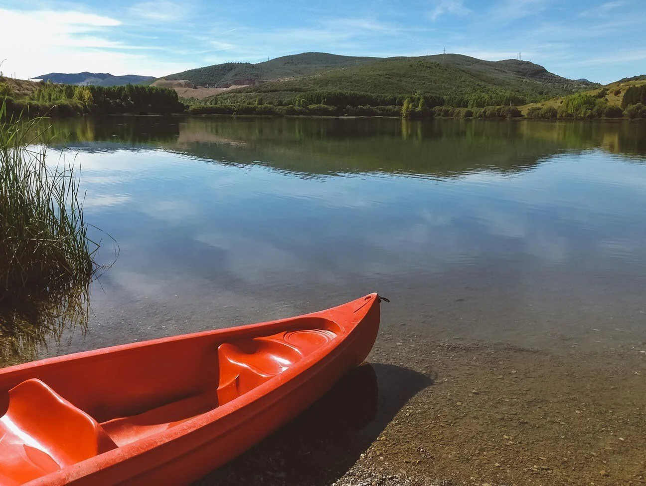 Un bote de kayak anaranjado en la orilla de un río rodeado de montañas y vegetación, con un cielo azul y algunas nubes.