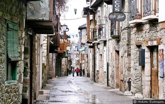 Calle peatonal en un pueblo antiguo con edificios de piedra y madera, bares y balcones, personas caminando.