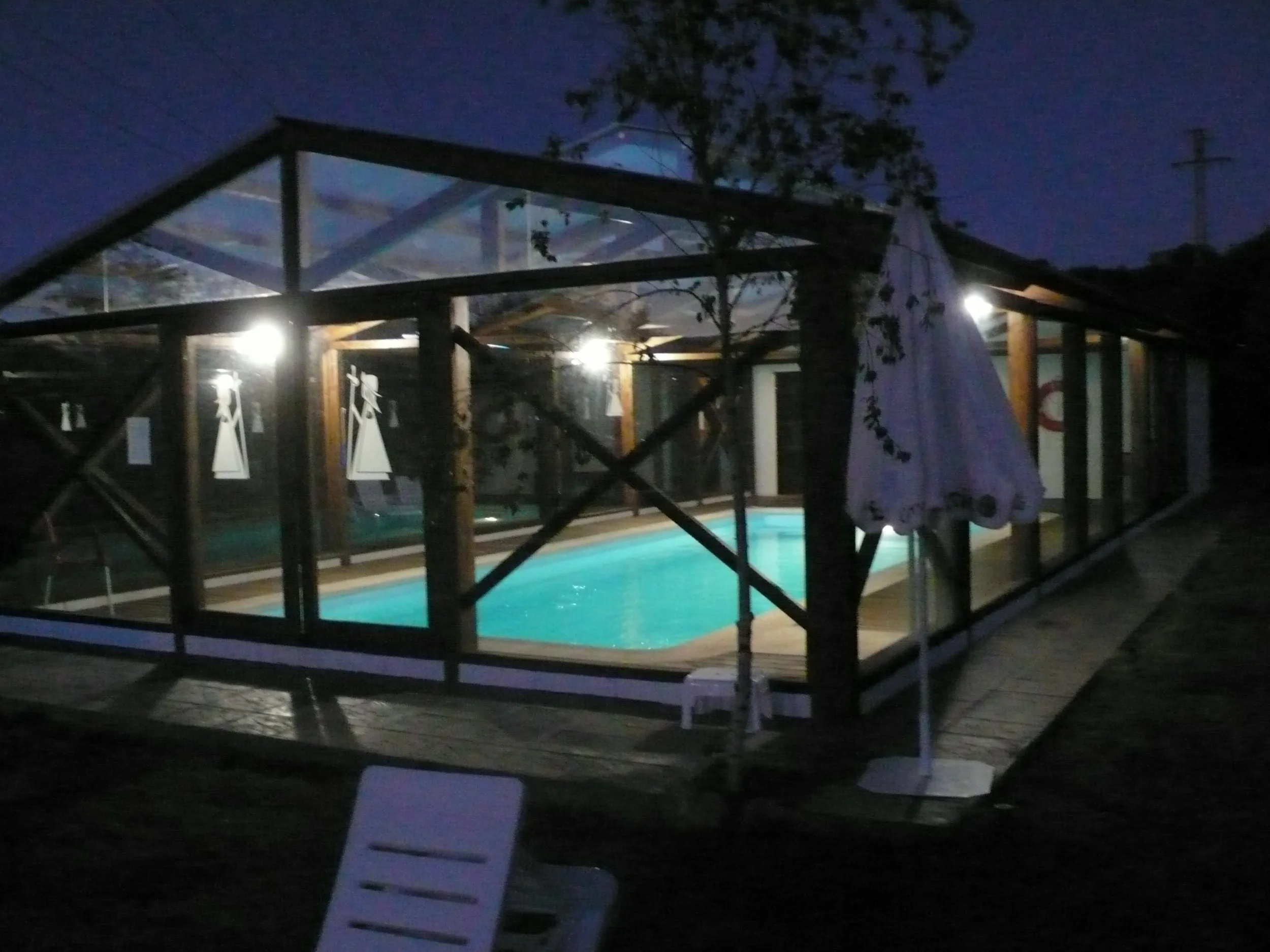 Piscina cubierta con estructura de madera y paredes de vidrio en un entorno oscuro, con iluminación interior y sombrillas en el exterior en hotel rural la peregrina