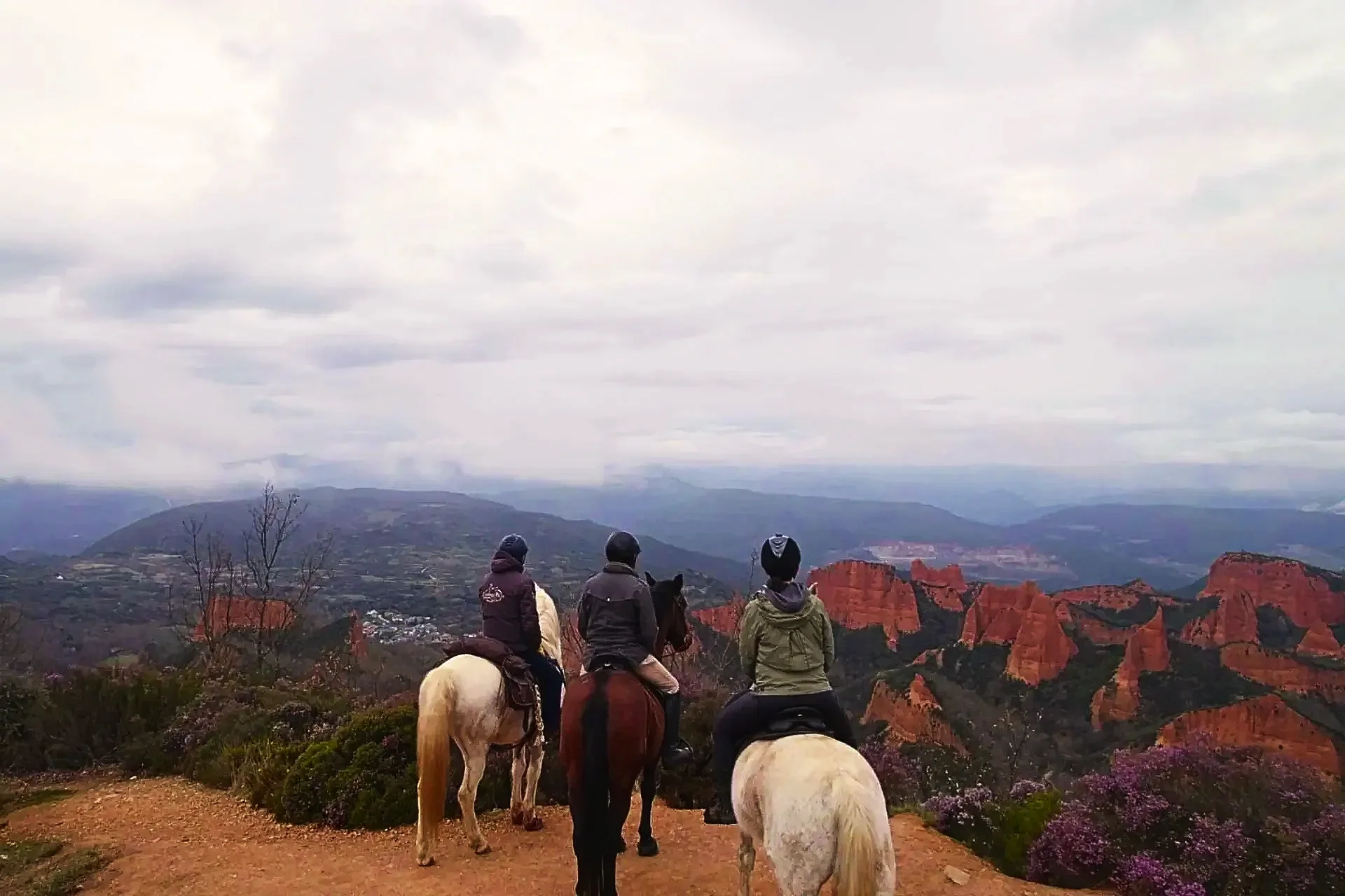 Tres personas montando caballos en un mirador, con formaciones rocosas rojas en el fondo bajo un cielo nublado.