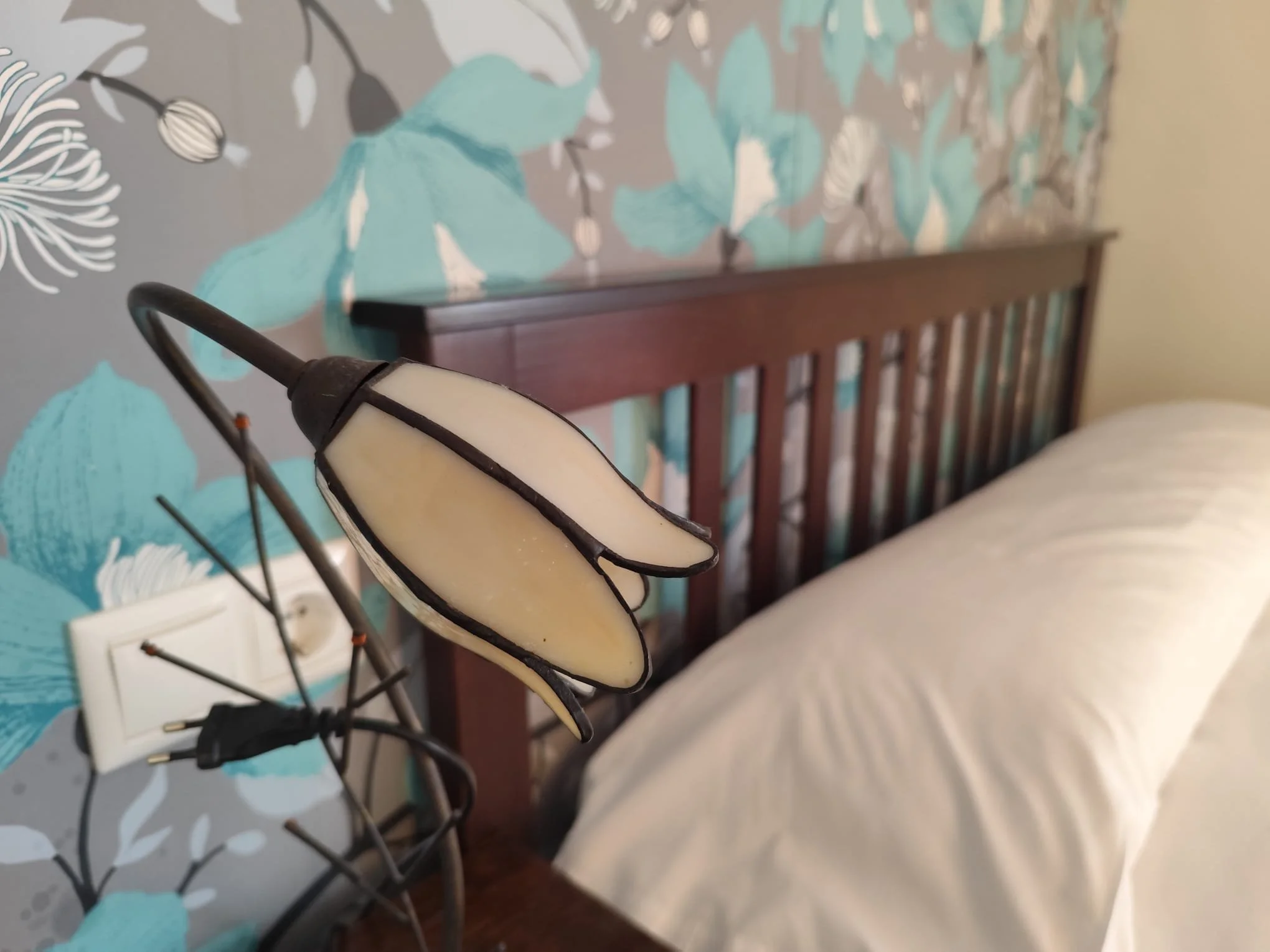 Lámpara de mesa con tulipa con forma de mariposa, en una habitación junto a la cama con cabecera de madera y papel tapiz con diseño floral en tonos azules y grisáceos en hotel rural la peregrina