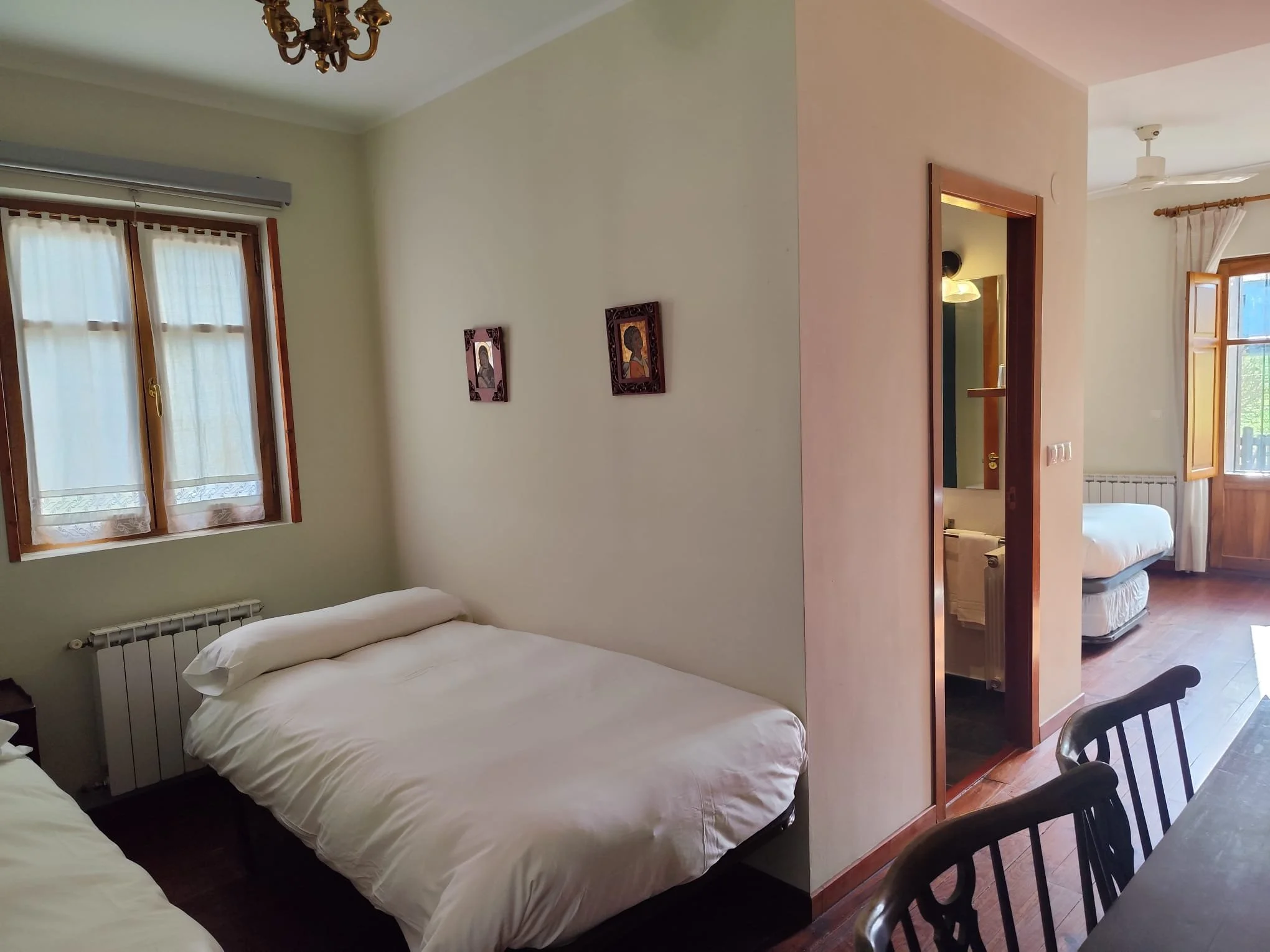 Habitación con dos camas, ventanas con cortinas, cuadros en la pared, baño visible desde la habitación, techo con ventilador y lámpara.