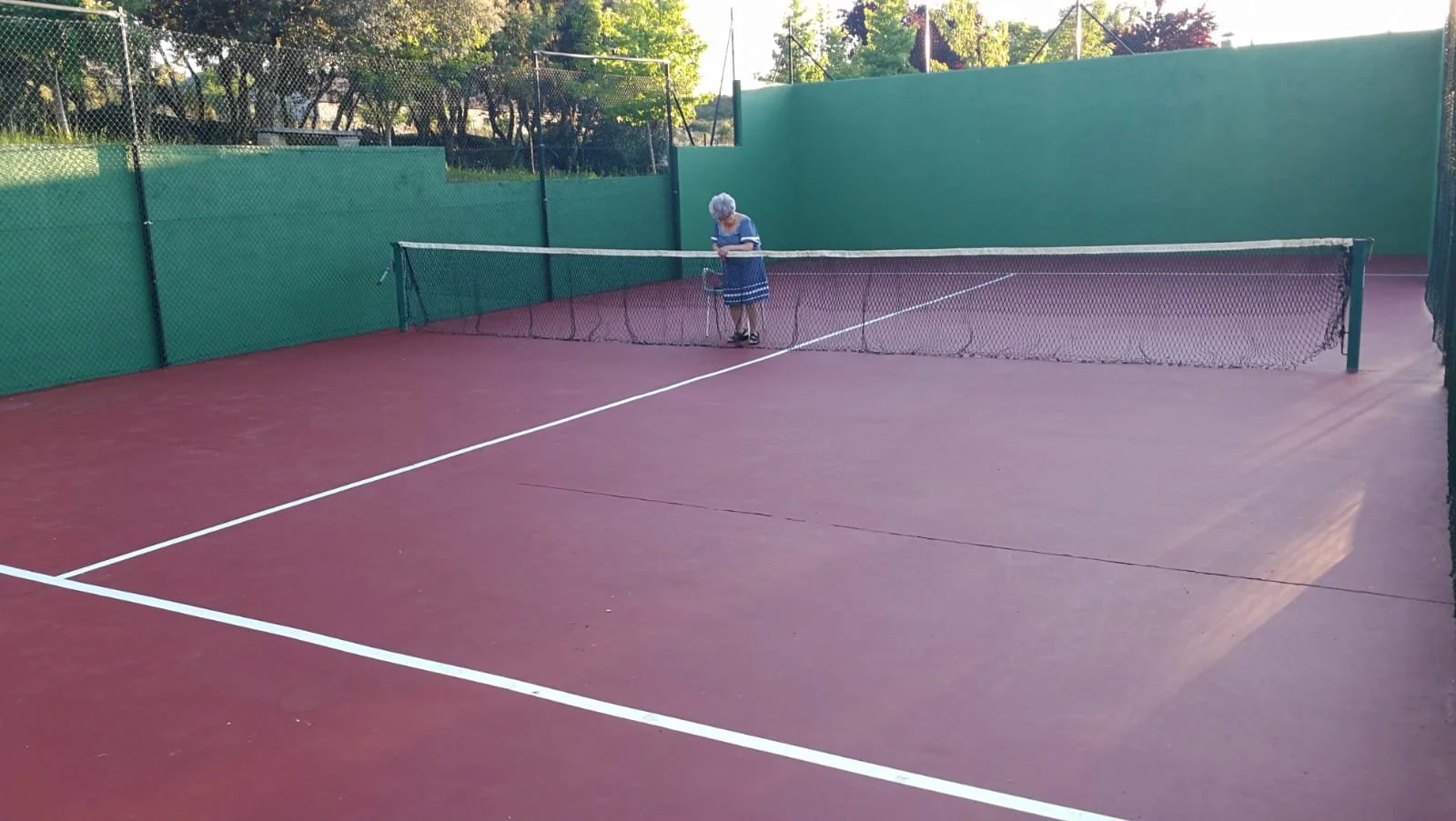 Pista de padel de tenis con una persona mayor vestida con un vestido azul, de espaldas, descansando en la red.