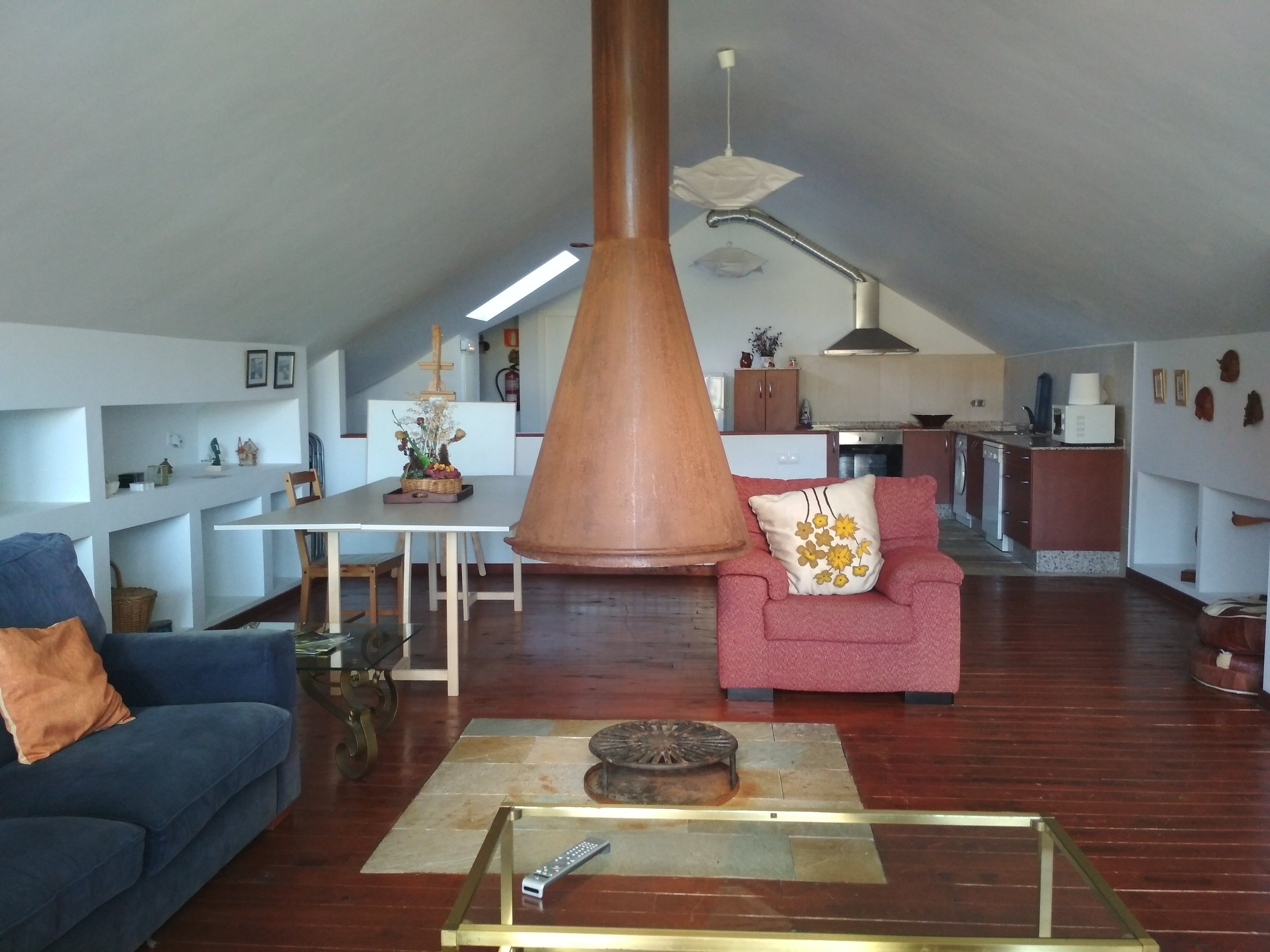Sala de estar con sillones, mesa de centro, cocina al fondo y decoración en paredes y repisas, en un espacio con techo inclinado.