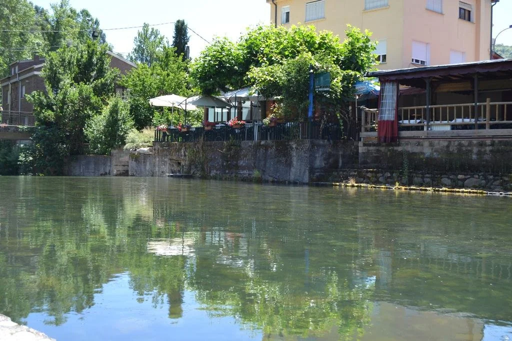 Vista de un río con agua clara, bordeado por una acera y casas con terrazas y árboles verdes en la orilla, en un día soleado.