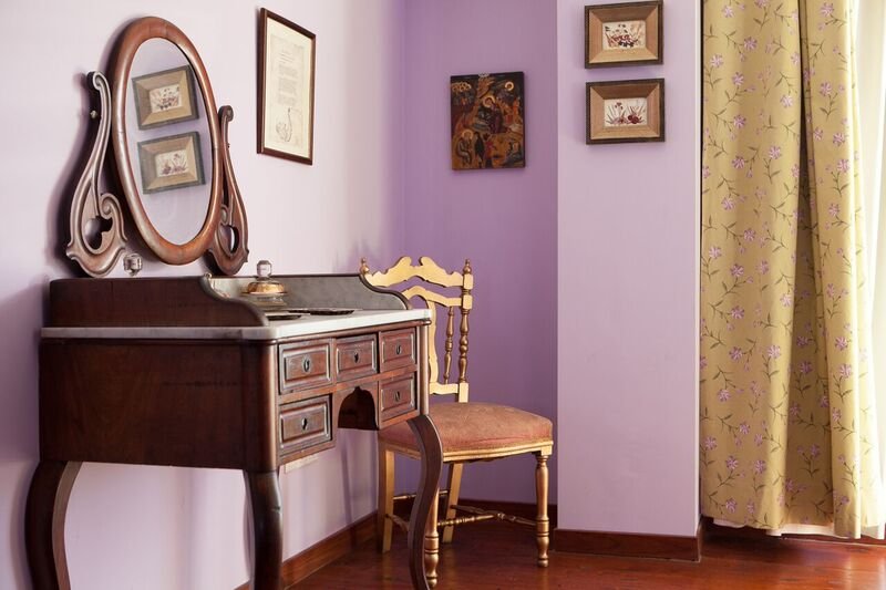 Rincón de una habitación con un tocador de madera, un espejo ovalado, una silla dorada con cojín y cortinas amarillas con borde floral. Hay cuadros en la pared de fondo.