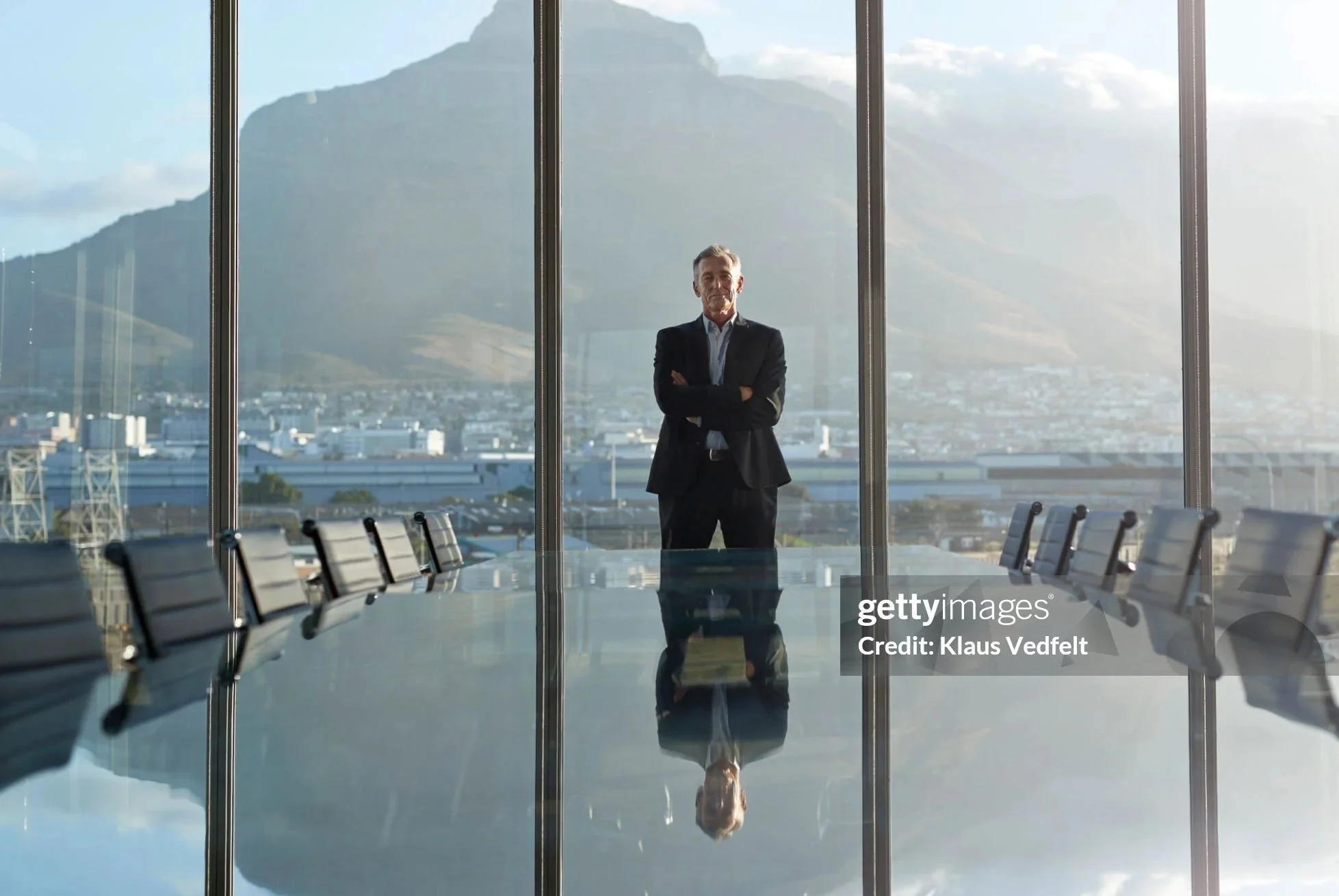 A businessman standing in a modern conference room with floor-to-ceiling windows, overlooking a city and mountain landscape during daytime.