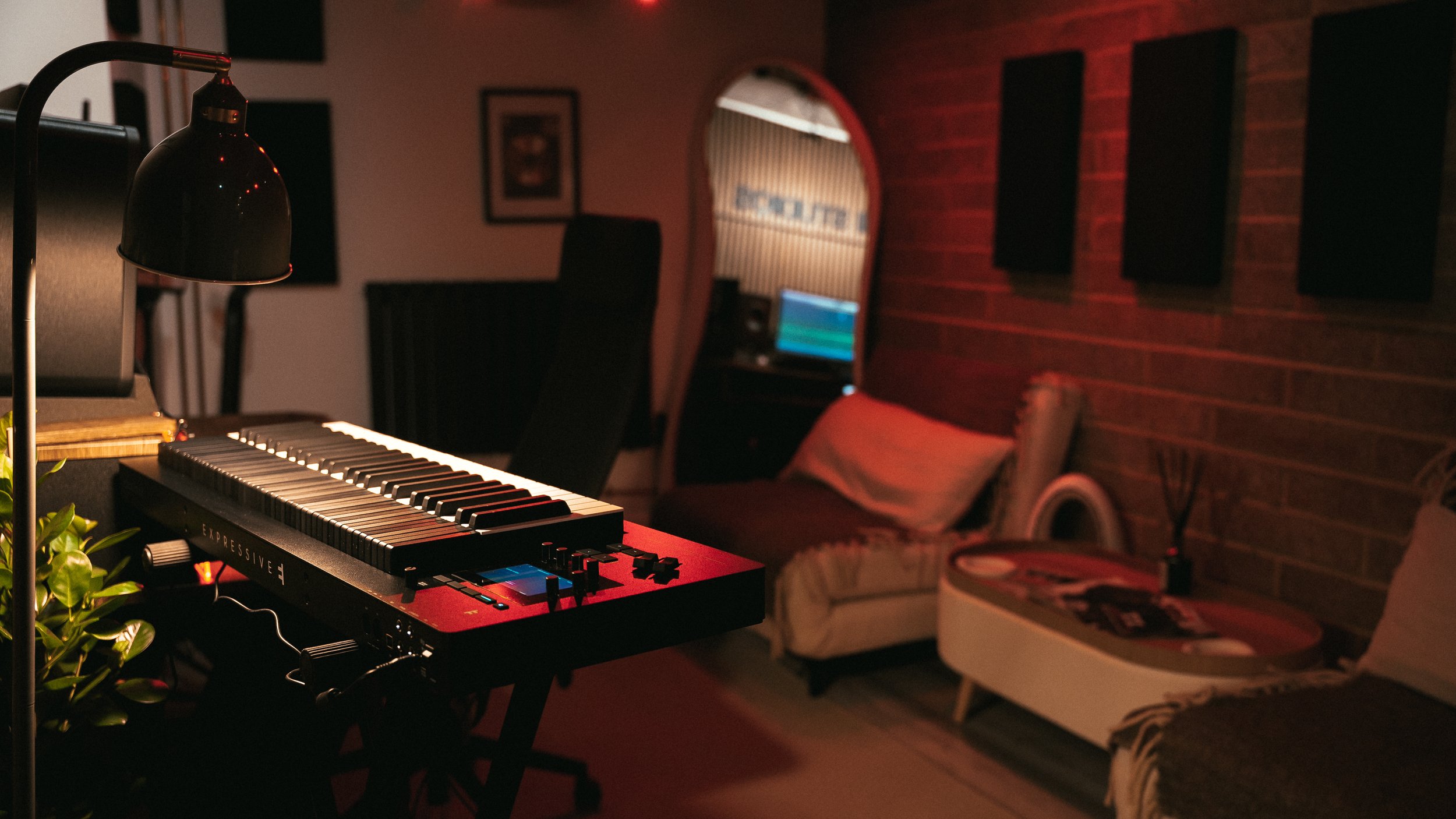 A music studio with a keyboard, a tabletop guitar, a lamp, and a television. The room has wooden walls, a cozy couch, and a small desk with a laptop.