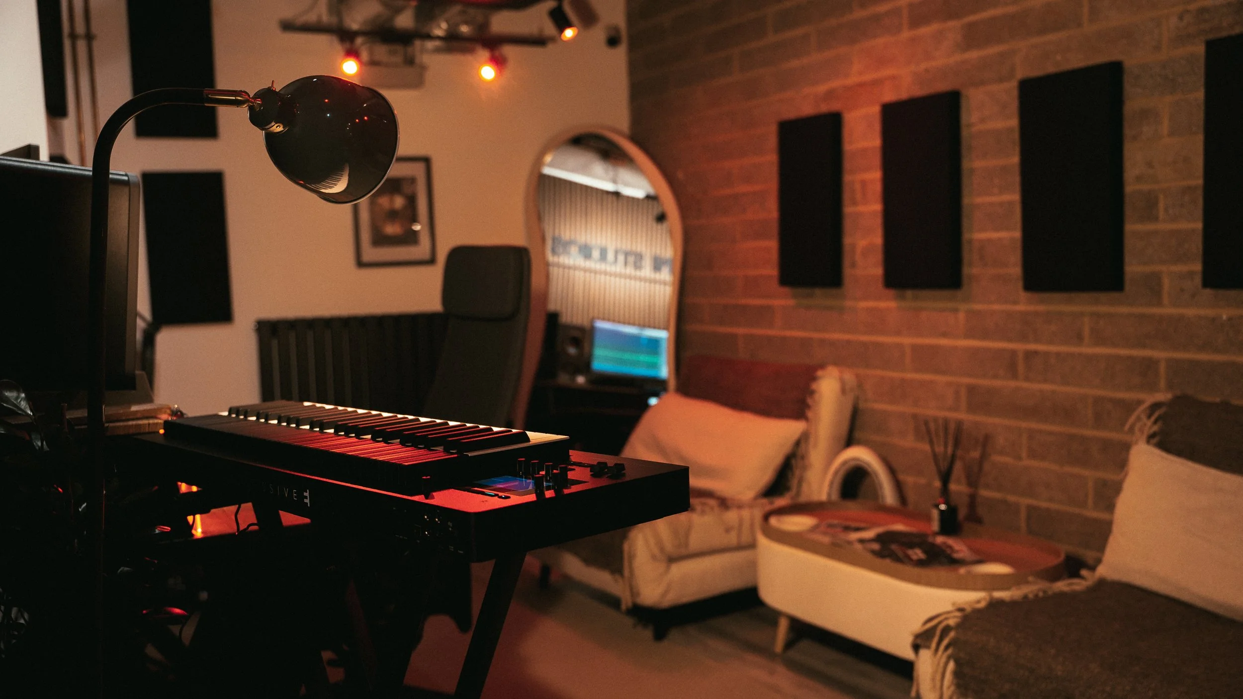 A music studio with a keyboard, computer monitor, and office chair in the foreground. There is a sofa, a side table with a diffuser and magazines, and a brick wall with black acoustic panels in the background.