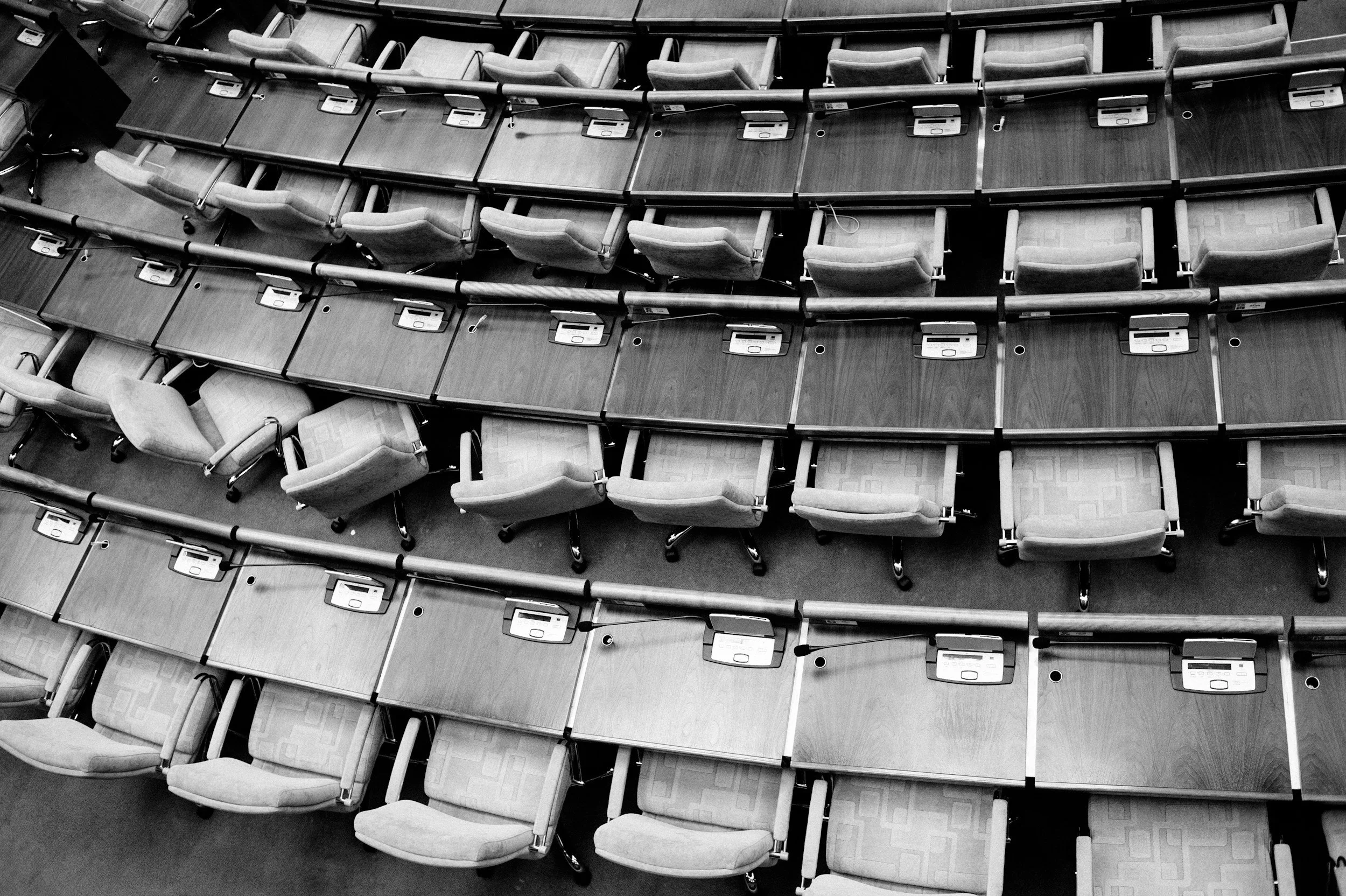 Empty rows of conference or legislative chairs with built-in microphones and nameplates, arranged in a tiered seating area, seen from above.