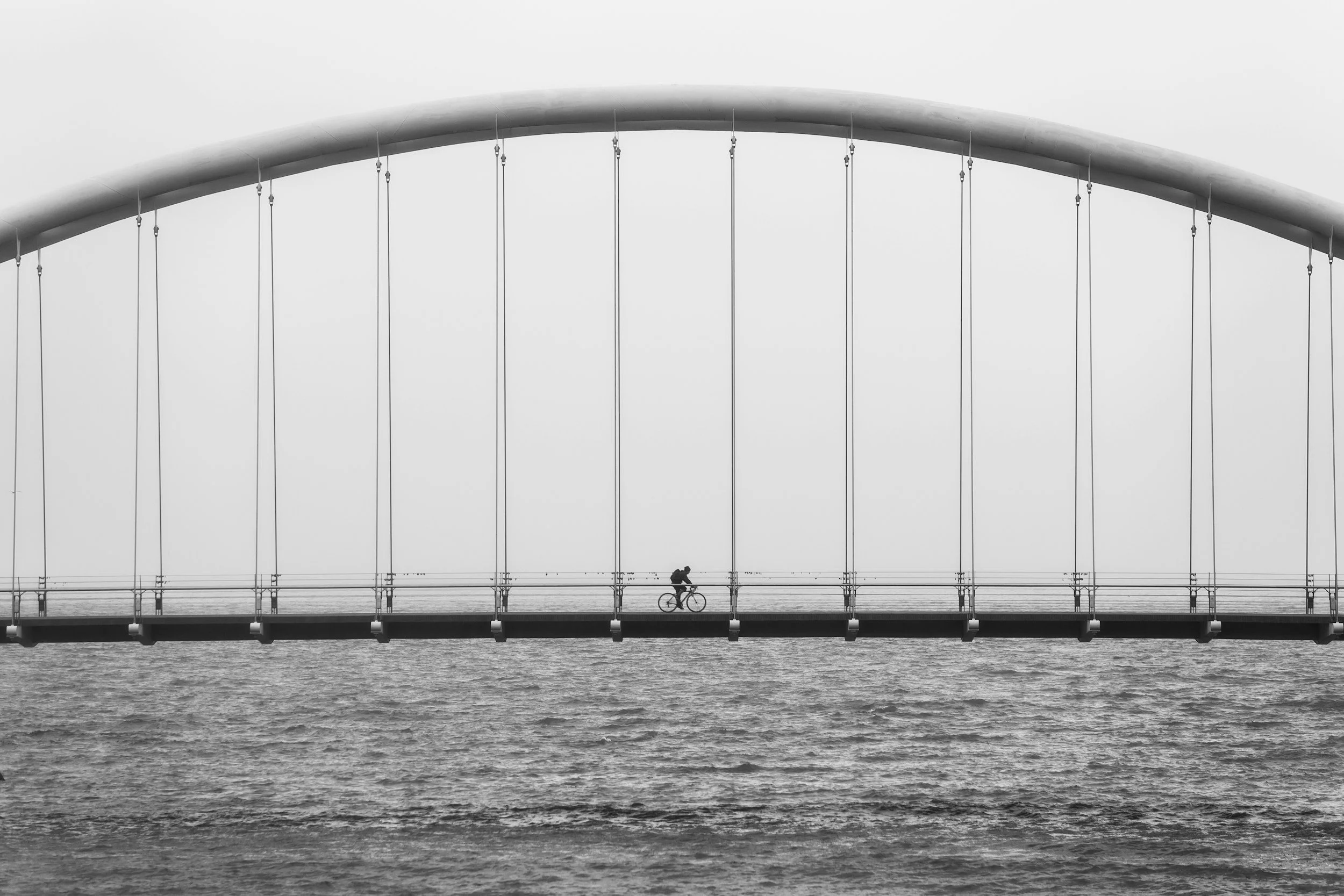 A black and white photo of a large suspension bridge over water with a single cyclist riding across.