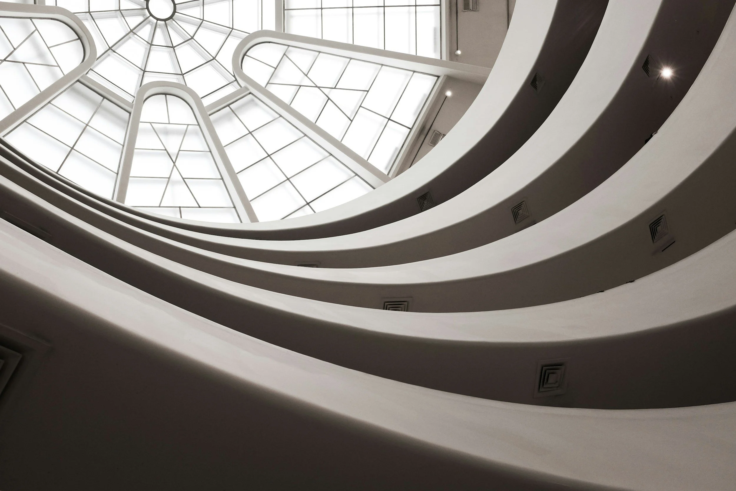Multiple white curved balcony railings in a building, viewed from below looking up towards a large glass ceiling.