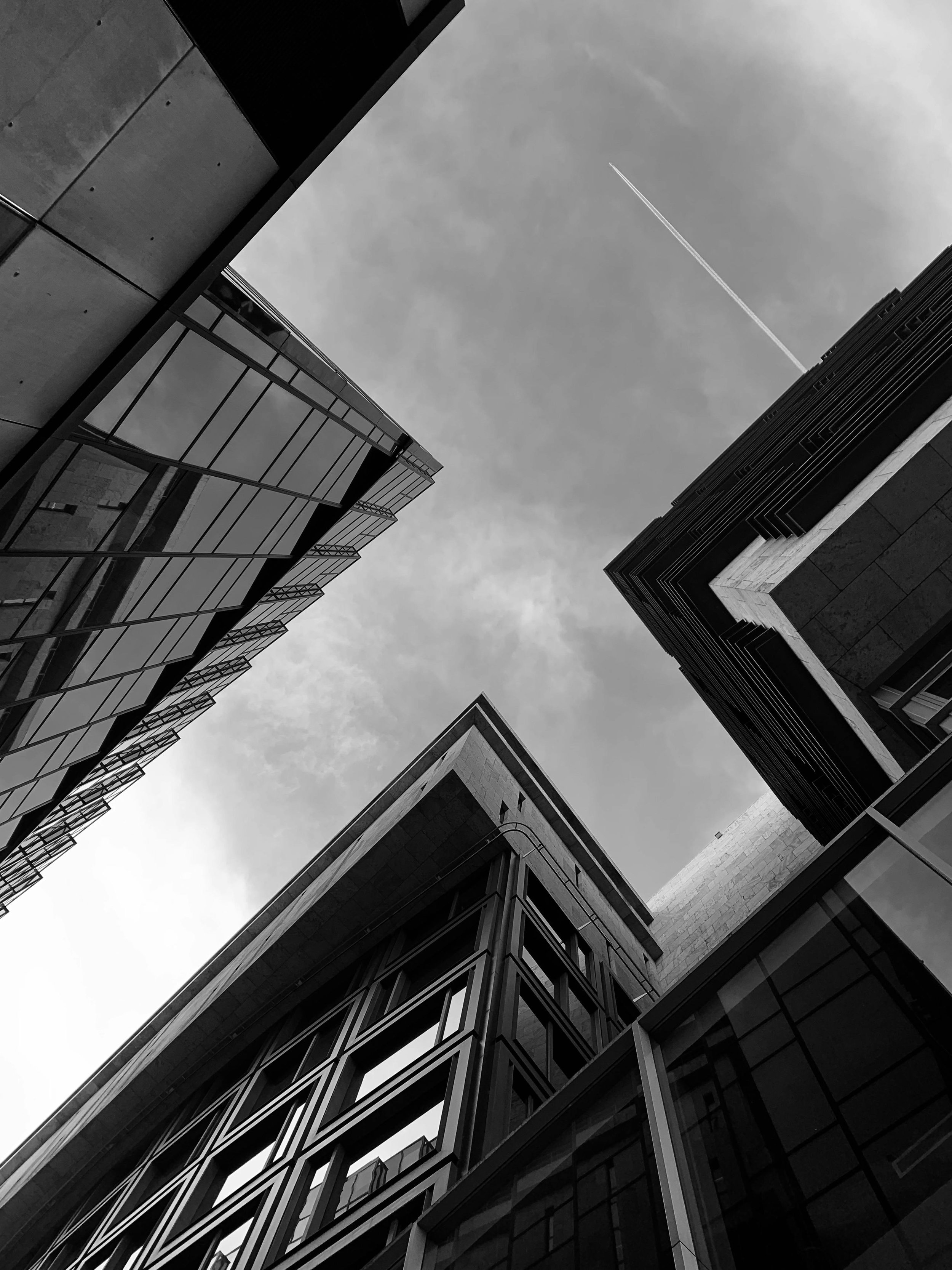 Black and white photograph of tall modern buildings taken from below, looking up at the sky with a contrail.