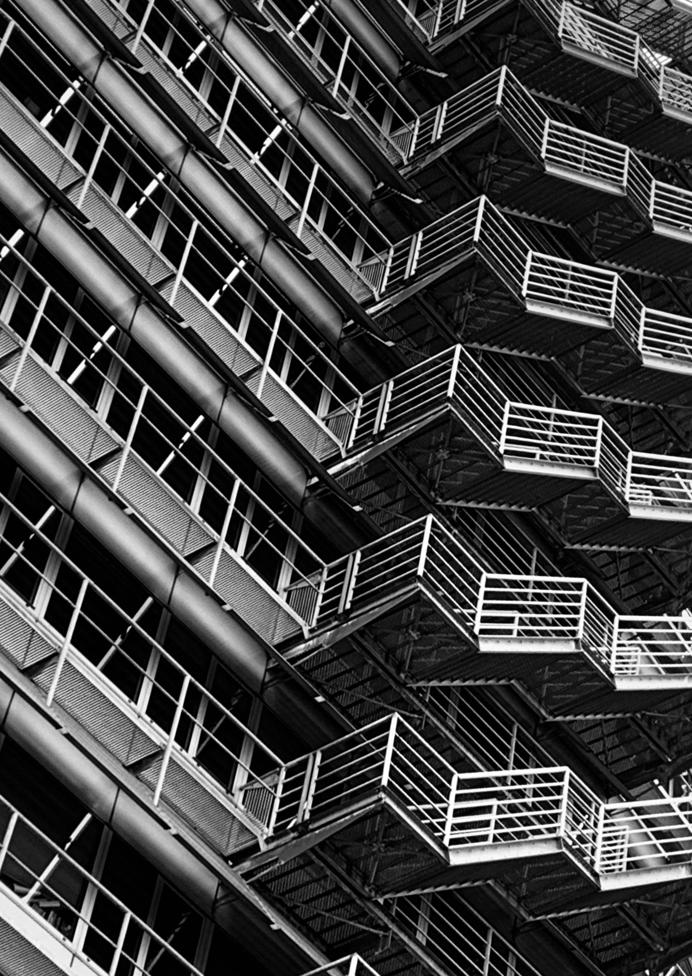 Black and white photo of an exterior building facade with metal fire escapes and balconies with railings, stacked diagonally across the building.