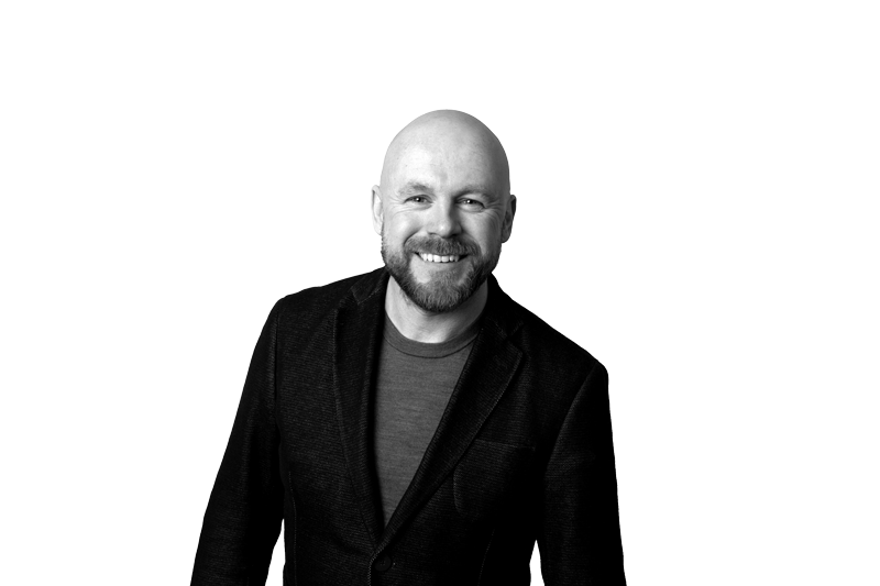 A smiling man with a beard wearing a dark blazer and a T-shirt, standing against a white background.