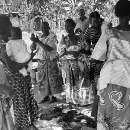 Group of women and children outdoors, some holding babies, on a patterned cloth with trees in the background.