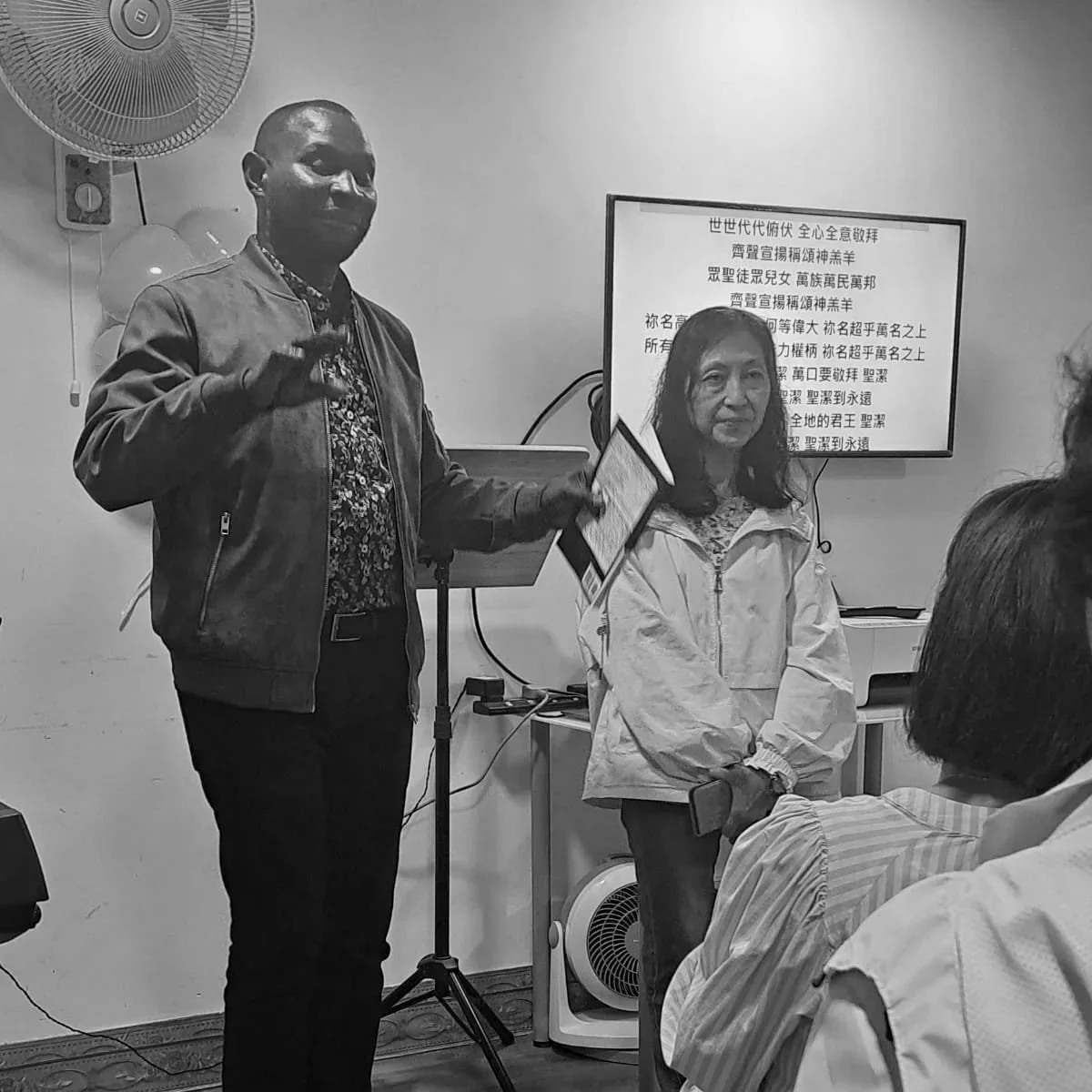 A black-and-white photo of two women and a man in a room. One woman is standing and holding a tablet, the man is next to her, gesturing with his hand. The other woman is sitting with her hands clasped, and a TV screen with Chinese characters is in the background. A fan is mounted on the wall.