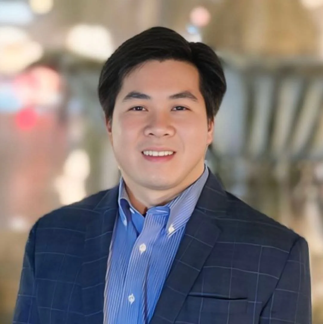A portrait of a young man with dark hair, wearing a checkered navy blazer and a blue collared shirt, smiling at the camera with a blurred cityscape background.