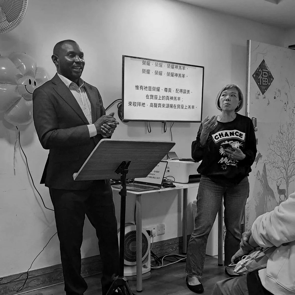 Black and white photo of a man in a suit and a woman in casual clothing speaking in front of an audience in a decorated room with balloons and wall art, with a screen displaying Chinese characters in the background.