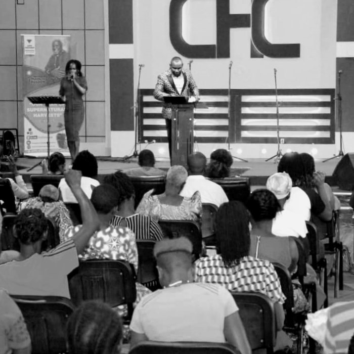 A man on stage giving a speech or presentation at an event with the CFC logo in the background, and an audience seated in front of him. A woman is standing to the side of the stage.