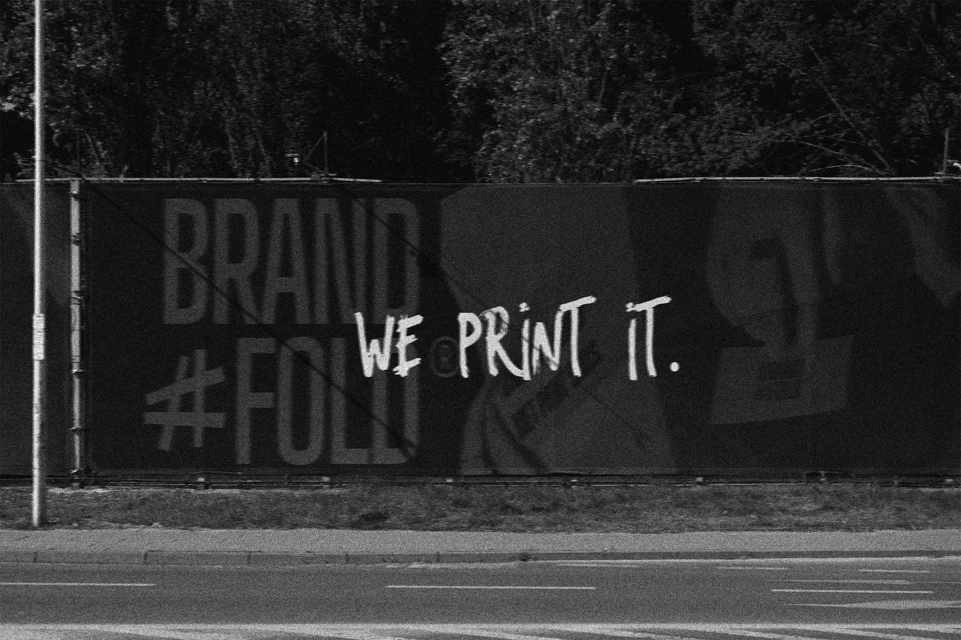 Hekwerk met graffiti en tekst 'We print it' in zwart-wit, in een buitenomgeving met bomen op de achtergrond.
