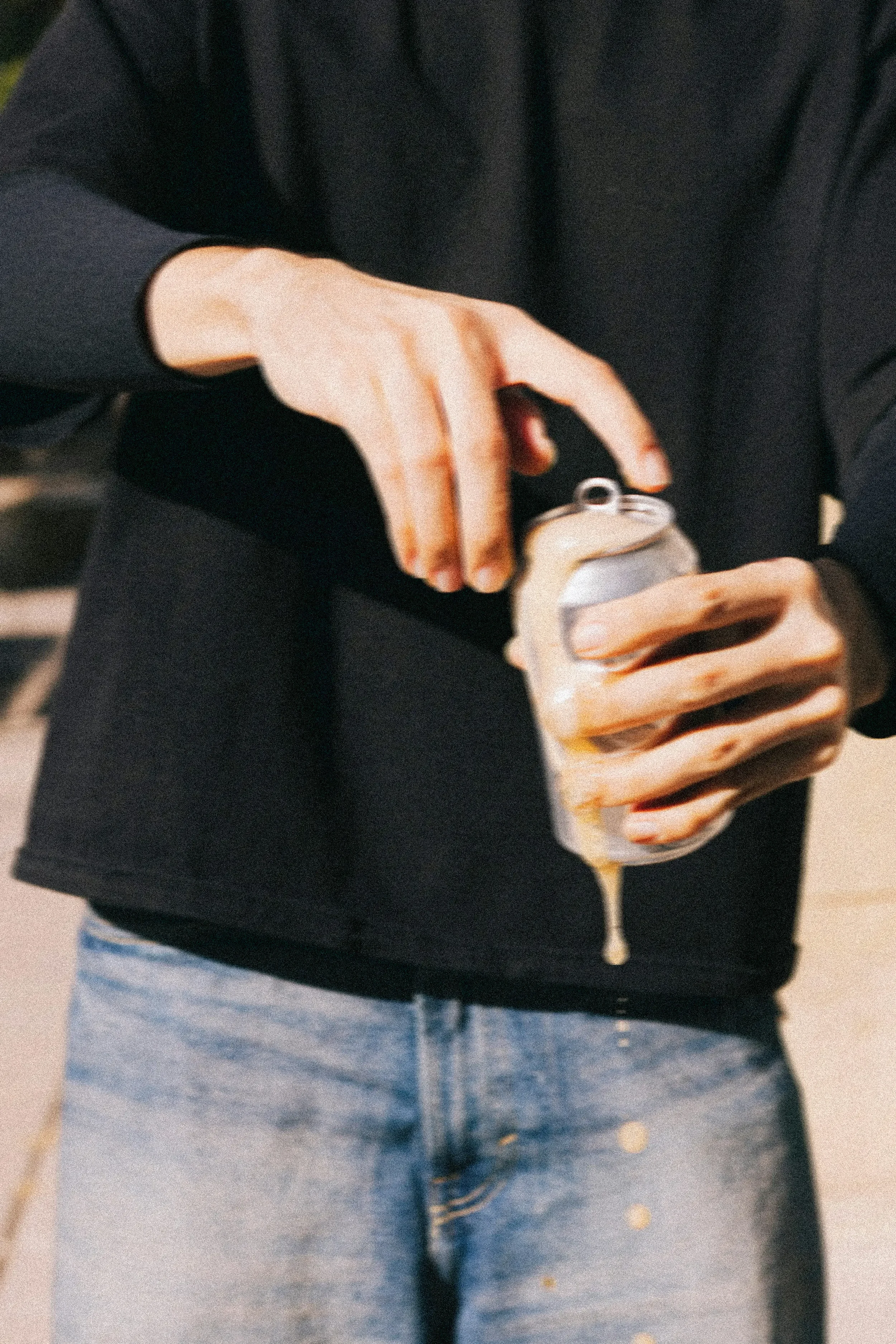 Persoon in een zwarte trui en blauwe spijkerbroek opent een blikje met een vinger en druipt honing uit het blikje.
