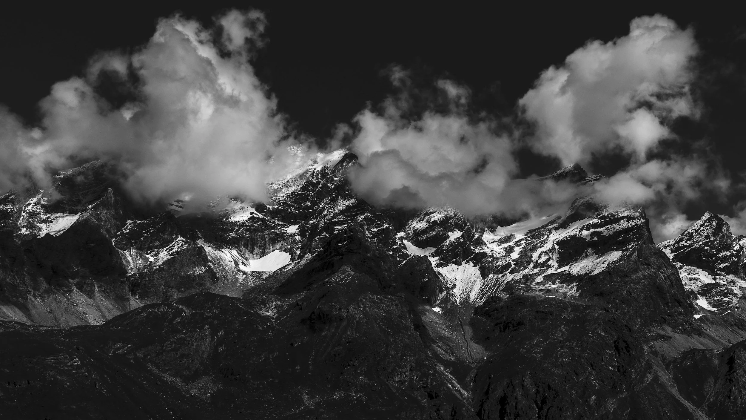 Black and white photo of mountain peaks with snow and clouds in the sky.