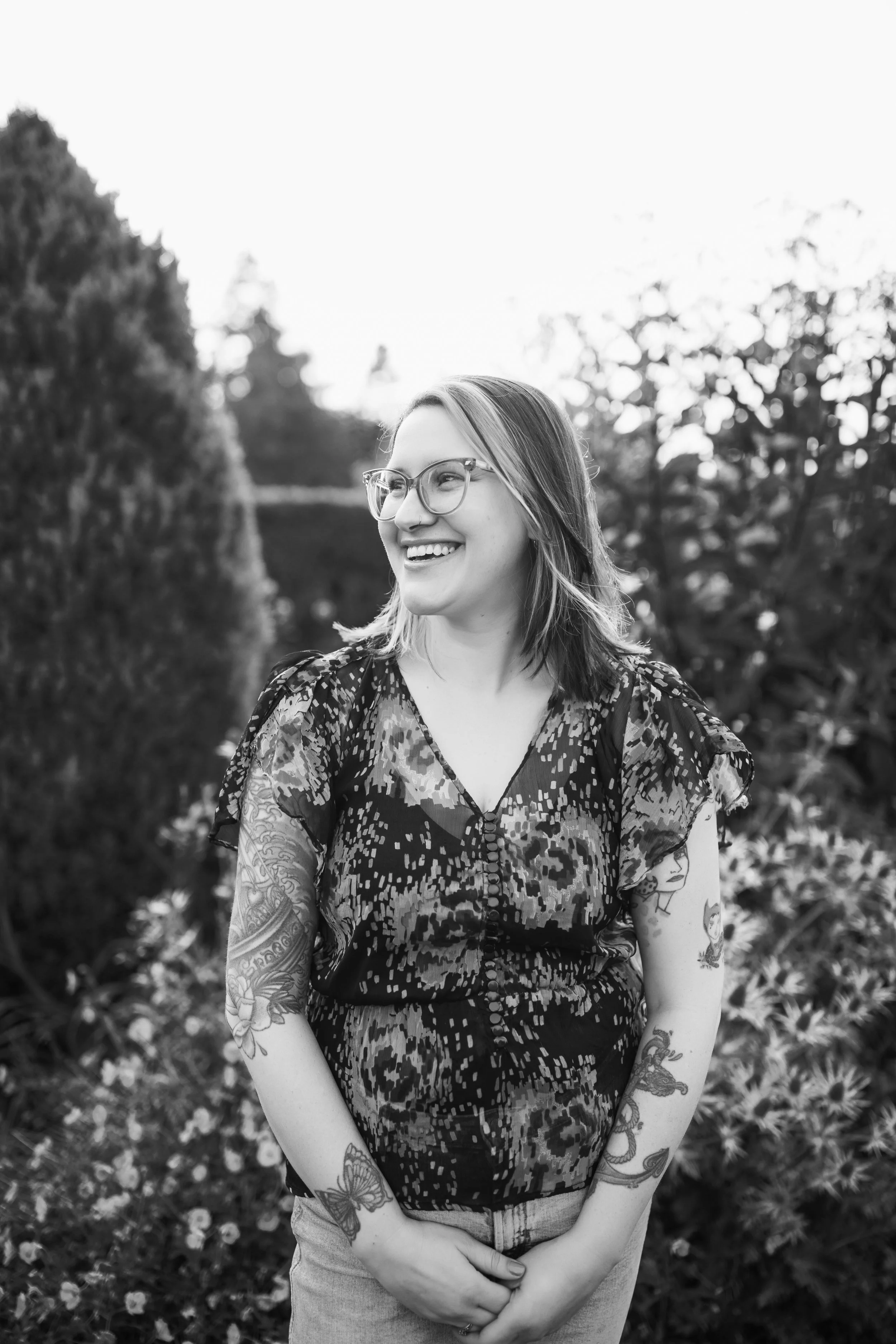 A smiling woman with glasses and tattoos on her arms standing outdoors among trees and bushes, captured in black and white.