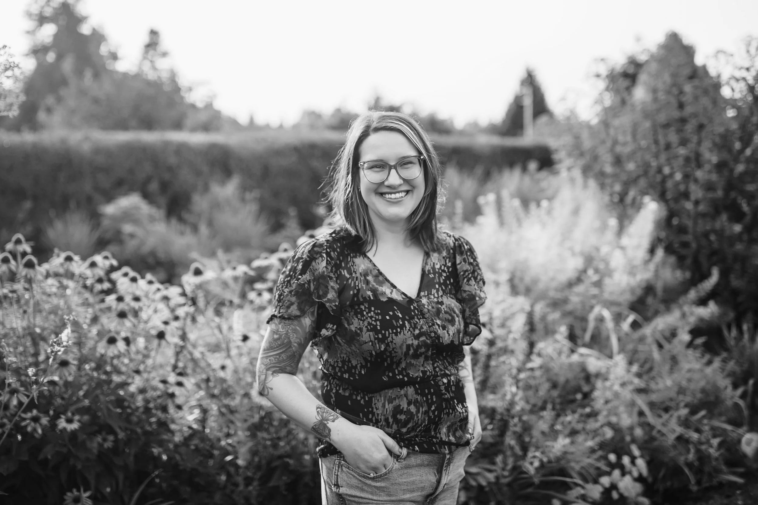 A woman with glasses and tattoos on her arms standing outdoors in a garden, smiling at the camera with a blurred background of plants and trees.