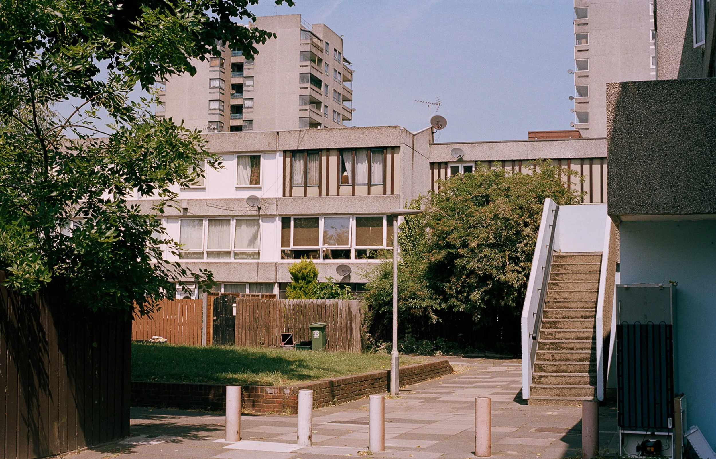 Residential apartment complex with trees, a staircase, a lamp post, and a small grassy area with a wooden fence and trash bins.
