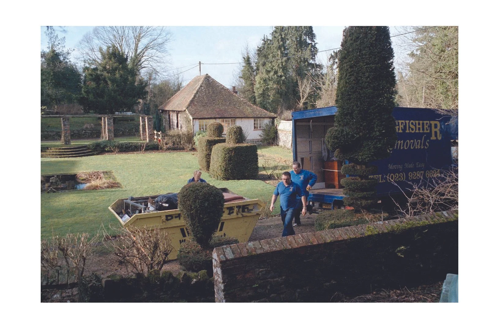 Movers in blue shirts unloading a moving truck in a garden with trimmed bushes and a small pond.