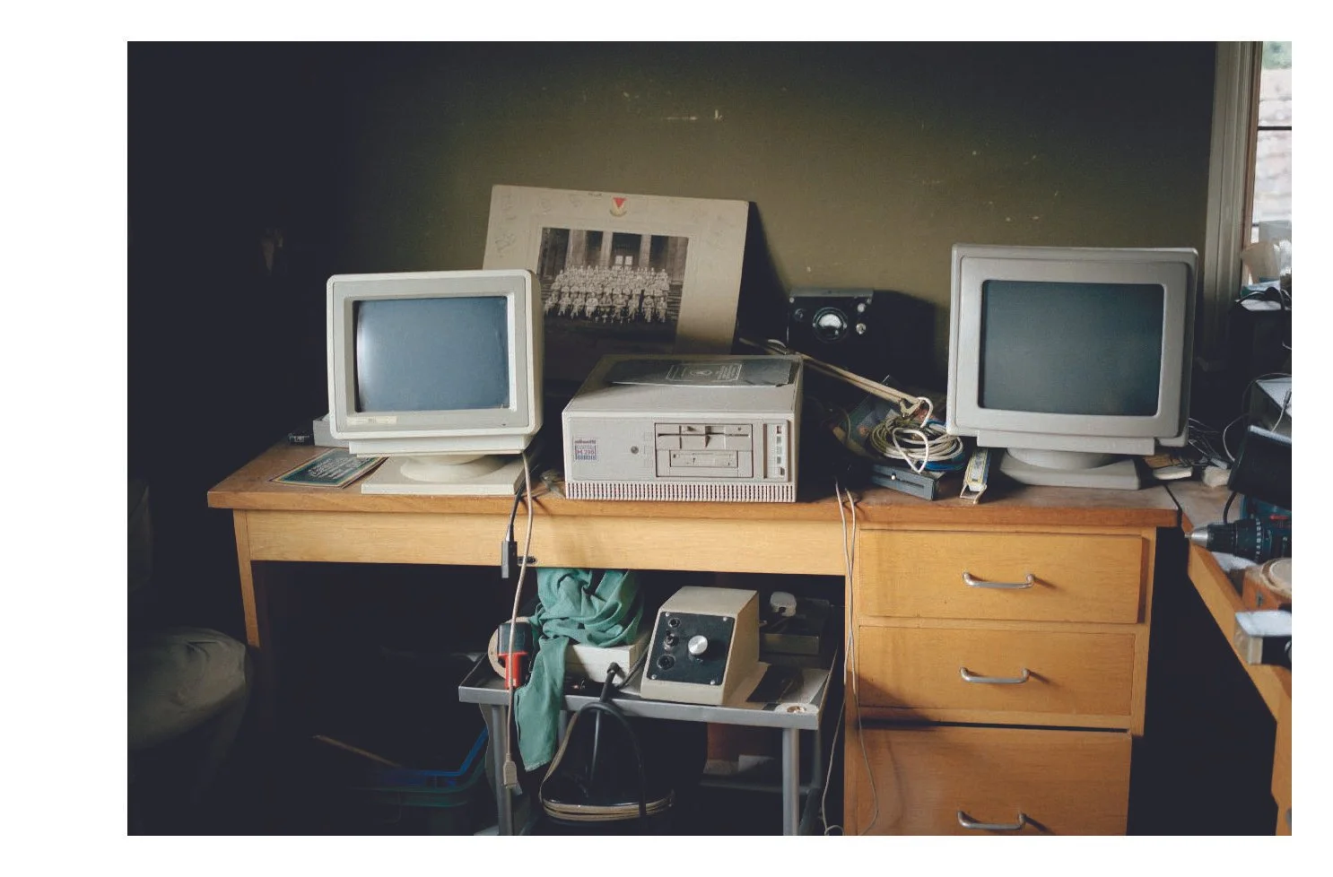 A cluttered wooden desk holding two older computer monitors, a large gray computer tower, a small speaker, and various cables. There is a wall behind the desk, a framed photo of a group in front of a building, and a window on the right side.