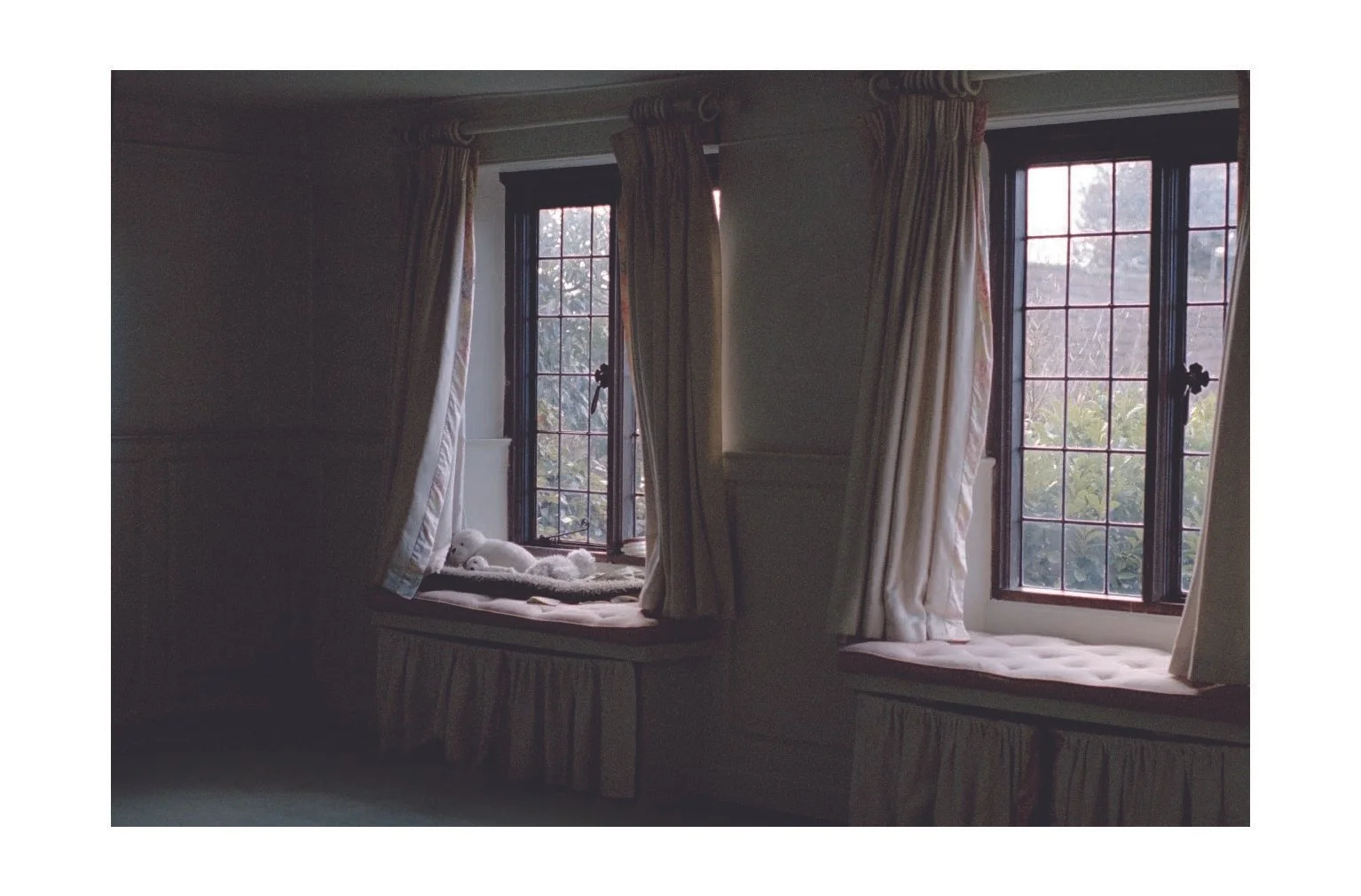 A cozy window seat with curtains and a stuffed animal, overlooking a garden outside.