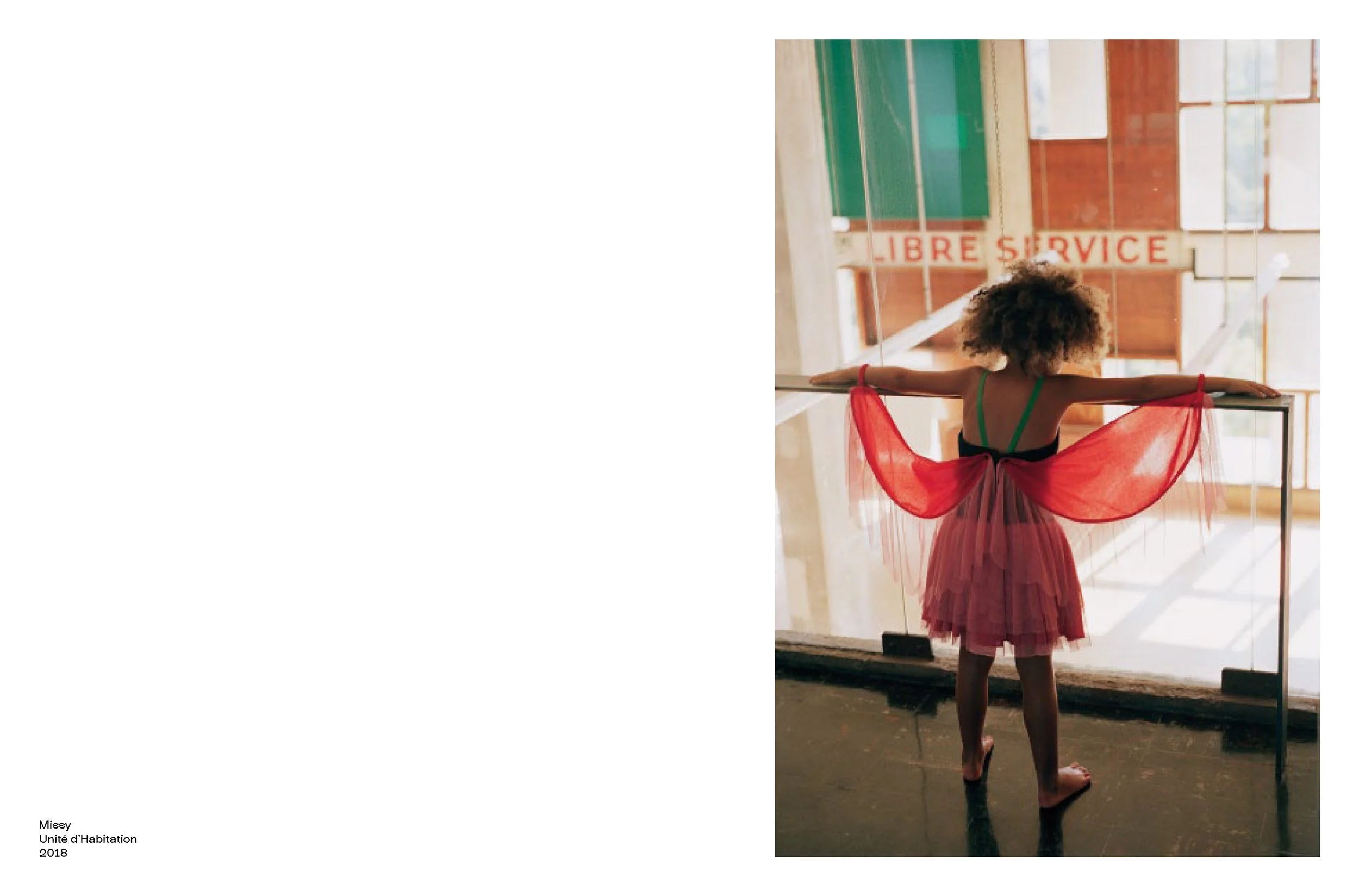 A young girl wearing a black top, green straps, and a pink tutu with red semi-transparent wings stands facing away at a glass window or door with her arms extended and holding onto the edge. Behind her, a sign reads 'Libre Service' in red letters. Th
