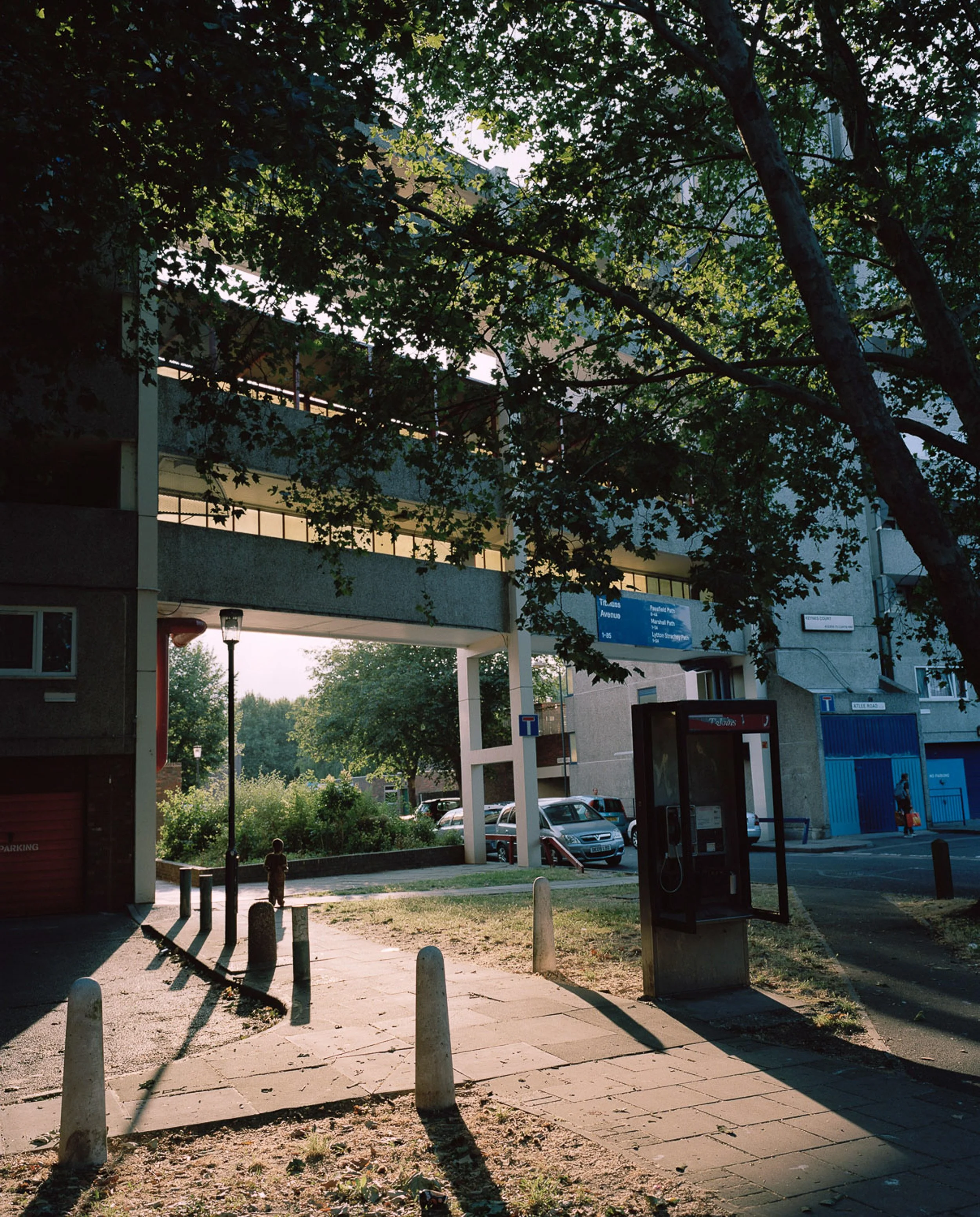 Street view showing an urban area with a multi-story residential building, parked cars, and trees. A child is walking along the sidewalk near a phone booth, with shadows cast by the setting sun.