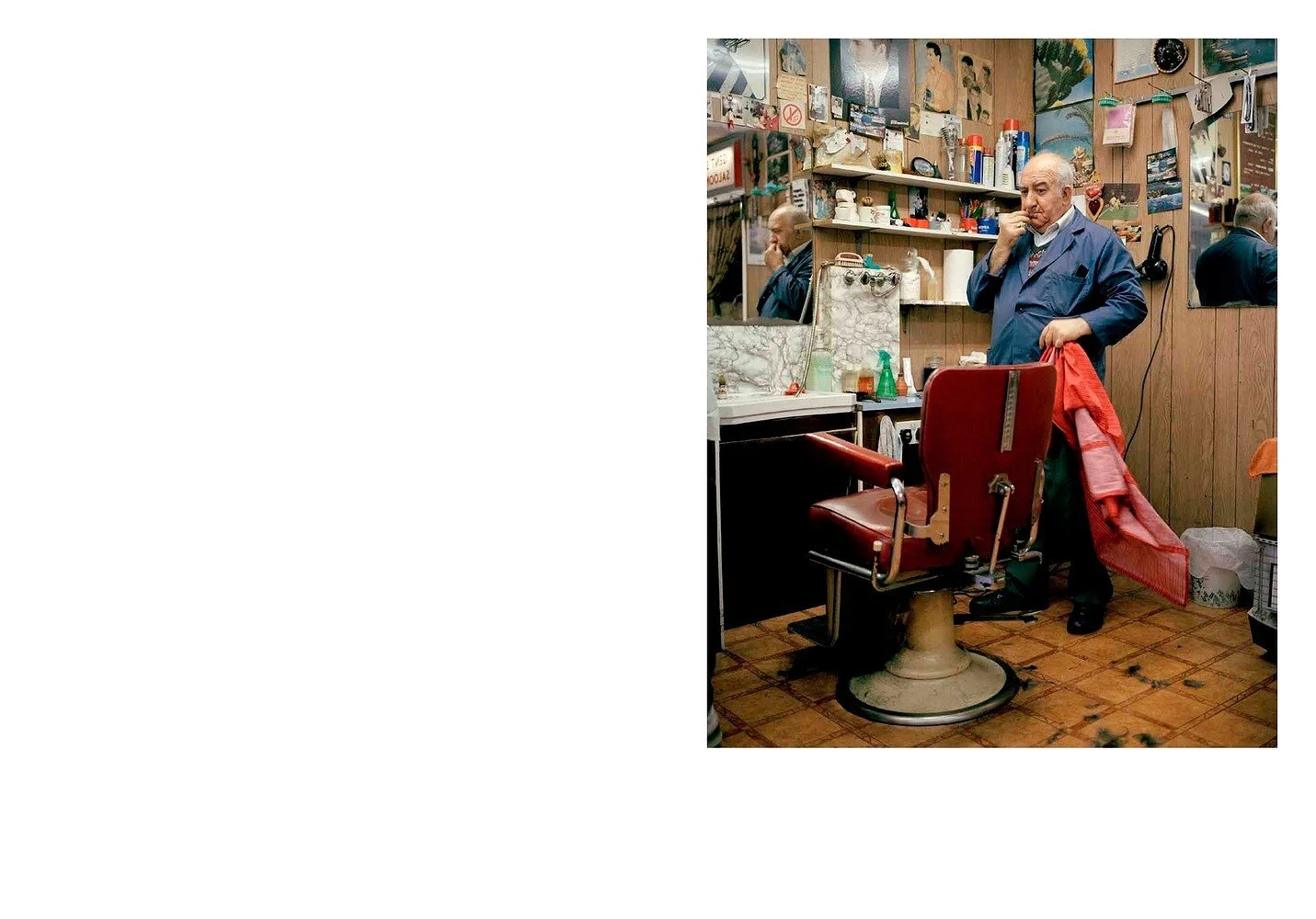 A barber shop with a barber holding a red cape, standing next to a vintage barber chair. The barber is thinking, with a hand on his chin. The walls are decorated with posters and various items.