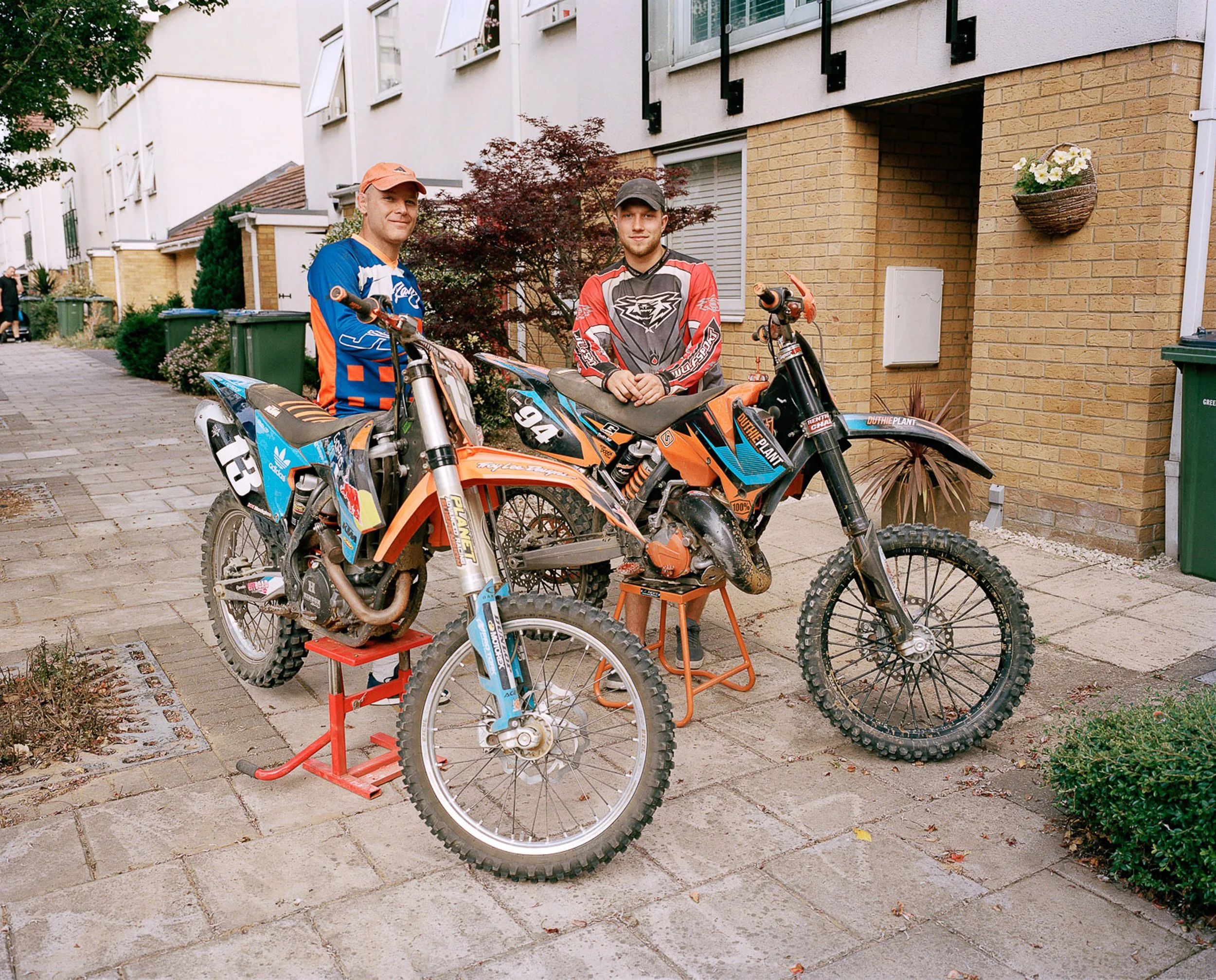 Two men in motocross gear standing next to their dirt bikes on a residential sidewalk.