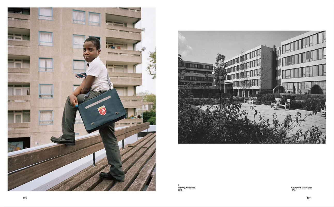 A young boy wearing a white shirt and gray pants sitting on a wooden bench outside a residential building, holding a navy school bag with a red crest