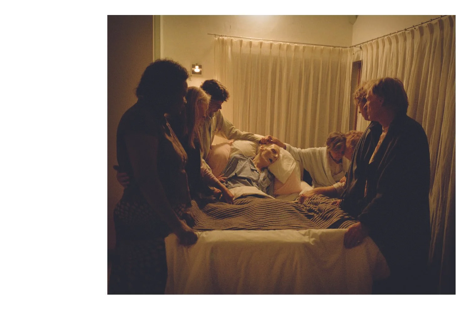 A group of six people and a dog gathered around a bed, with some holding hands and others resting on the bed, in a dimly lit room with a beige curtain and a small light on the wall.