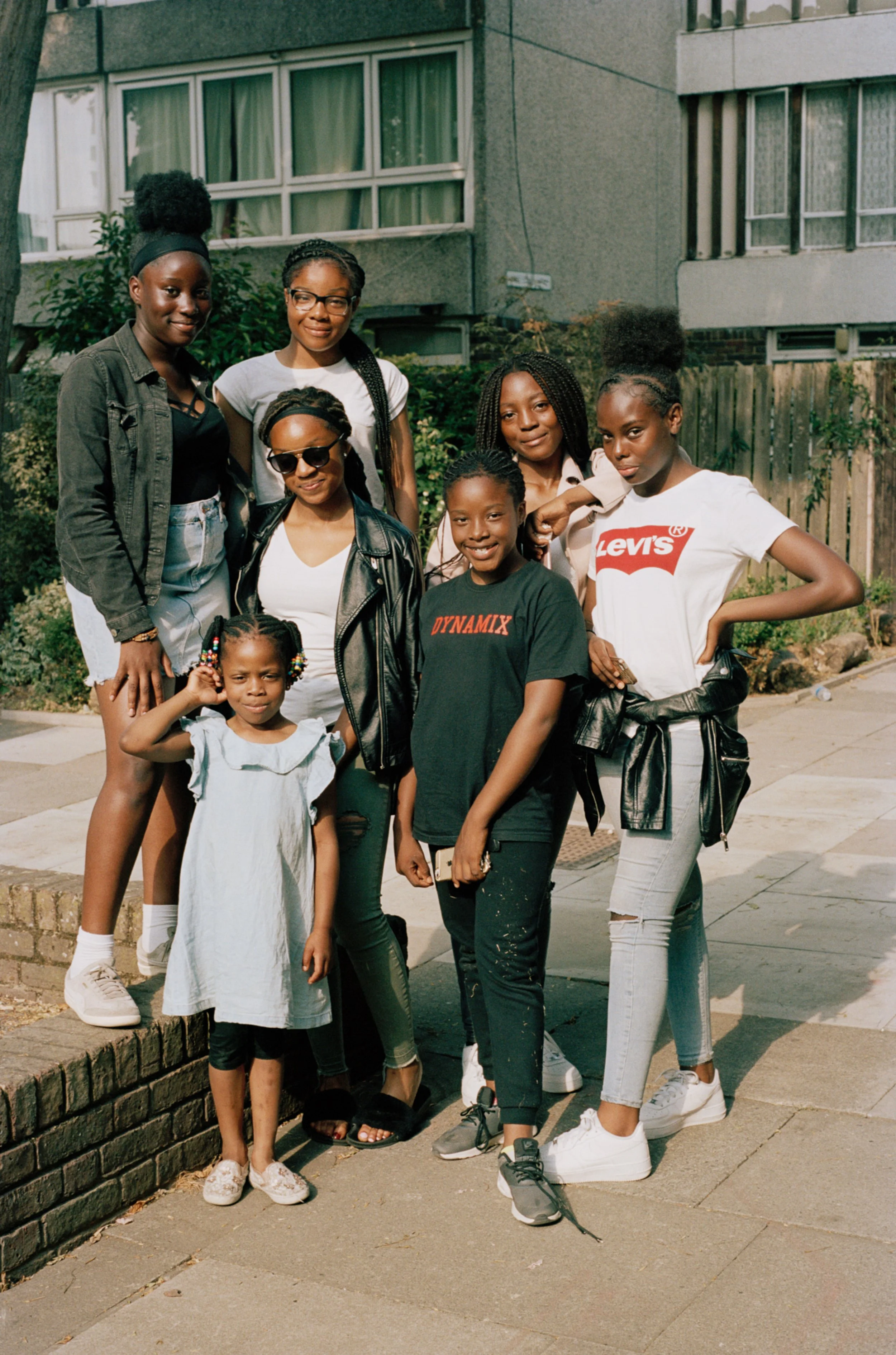 Group of eight girls of different ages standing together outdoors near a residential building and greenery, smiling at the camera.